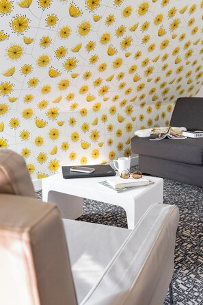 Um quarto com papel de parede estampado, uma mesa de centro branca com uma revista, um smartphone e uma xícara.