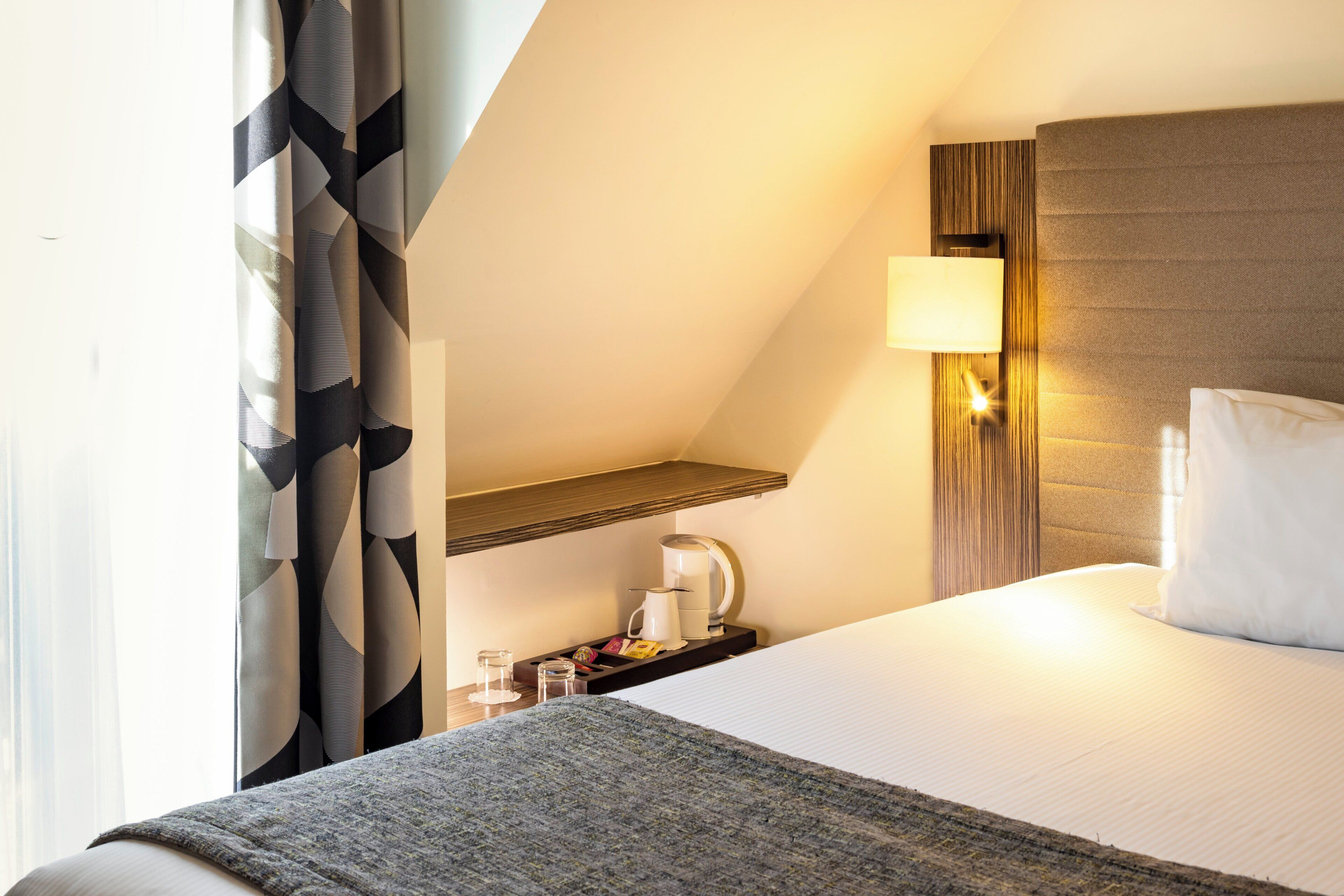 Une chambre d’hôtel avec un lit, une table de chevet, une lampe et une fenêtre avec des rideaux.
