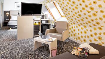 A hotel room with a bed, a TV, a desk with a laptop, a chair, a sofa, and a patterned wall.