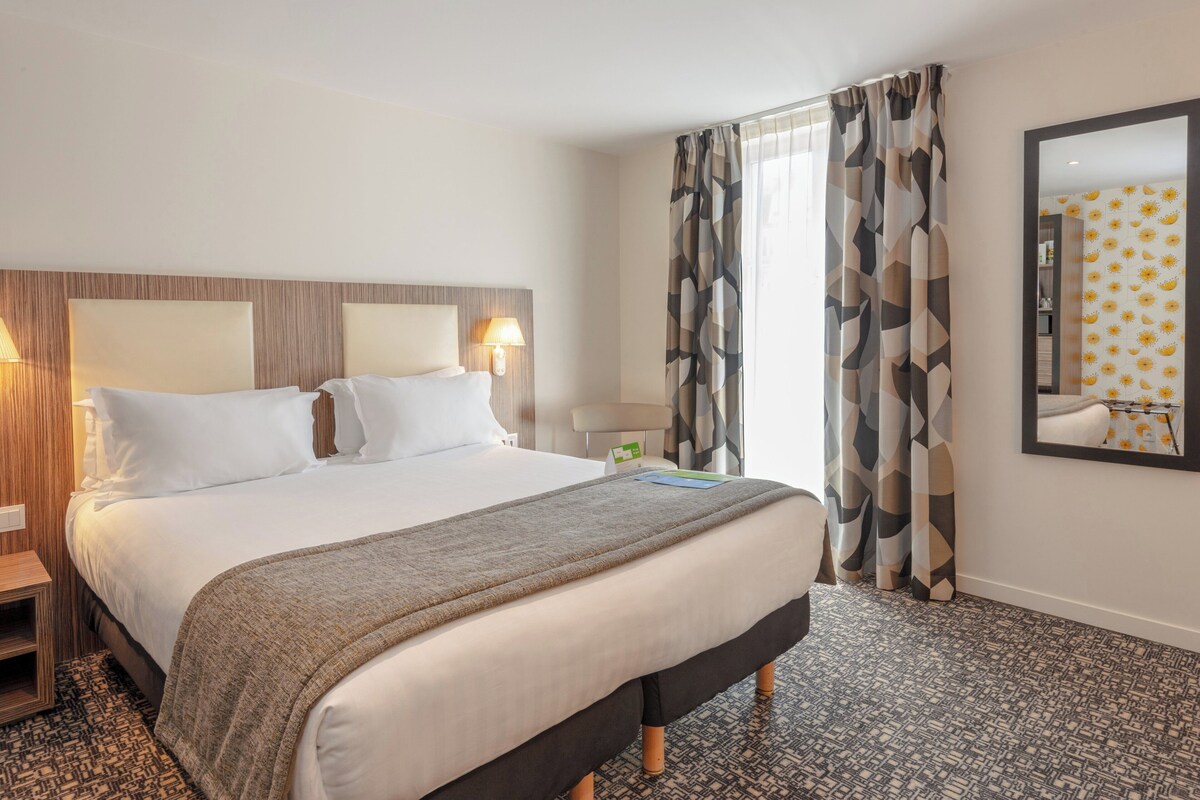 Quarto de hotel com uma cama grande, cabeceira de madeira, mesa de cabeceira com abajur, cortinas estampadas e um espelho na parede.