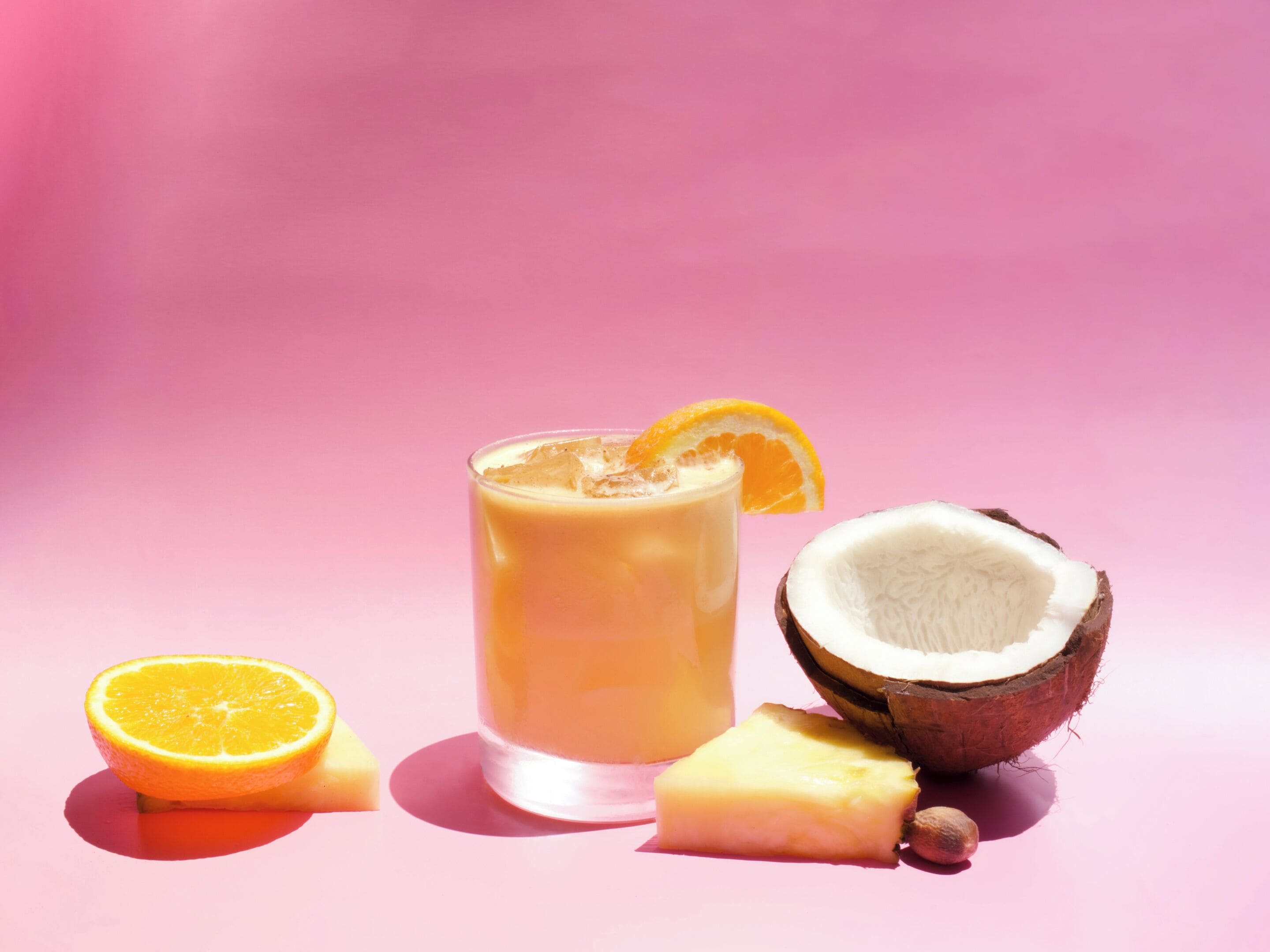 Ein Glas Orangensaft mit Orangenscheibe am Rand, eine halbe Kokosnuss und ein Ananasstück vor rosafarbenem Hintergrund.