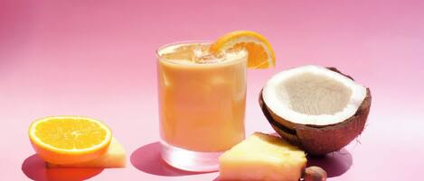 Ein Glas Orangensaft mit Orangenscheibe am Rand, eine halbe Kokosnuss und ein Ananasstück vor rosafarbenem Hintergrund.