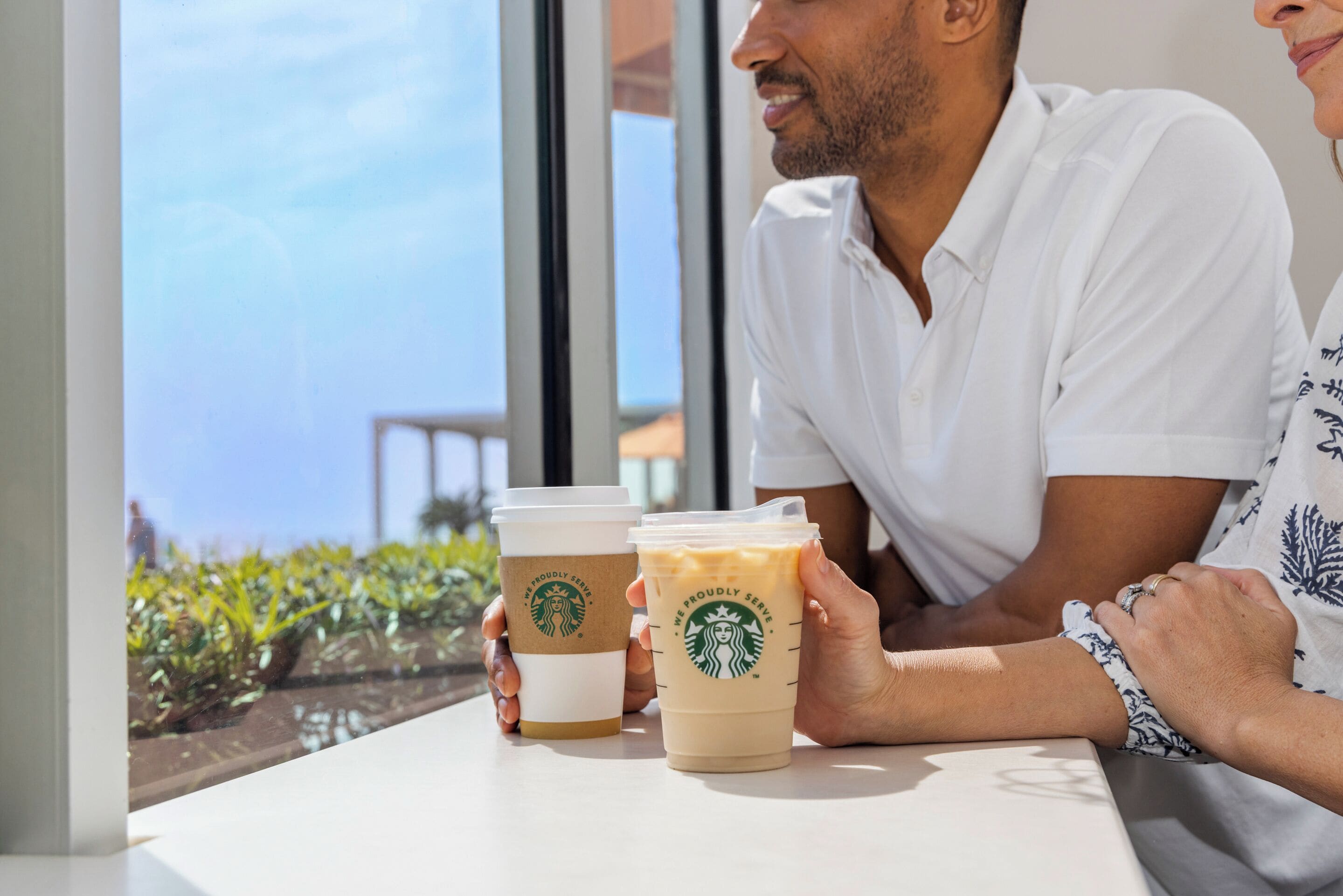 Eine Person hält zwei Starbucks-Tassen, im Hintergrund Grünflächen und ein Gebäude.