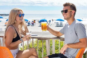 Deux personnes sont assises sur une terrasse en bois, d’où l’on a une vue sur la plage et l’océan.