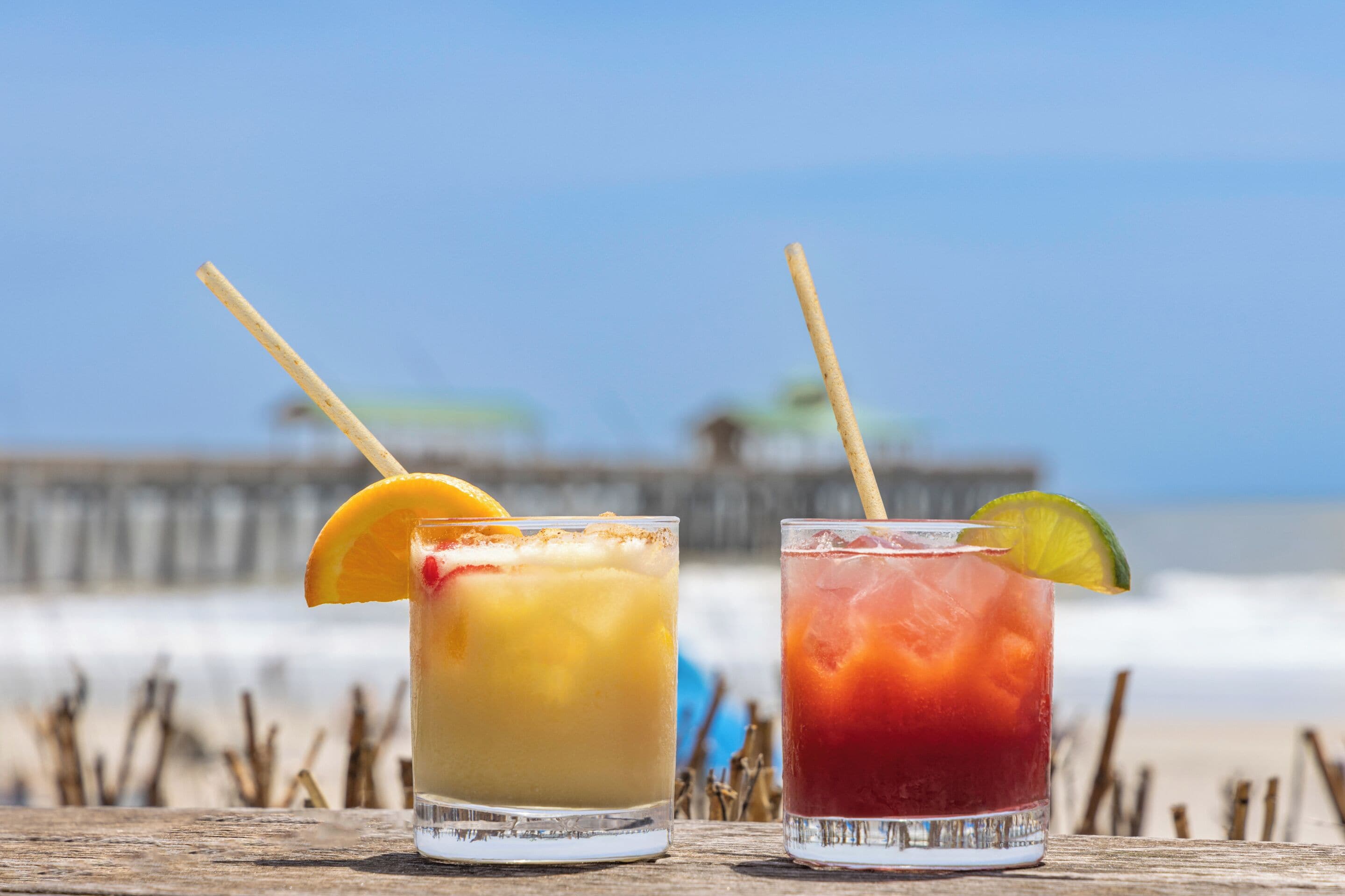 Dos vasos de bebidas heladas sobre una superficie de madera, con una playa y muelle al fondo.