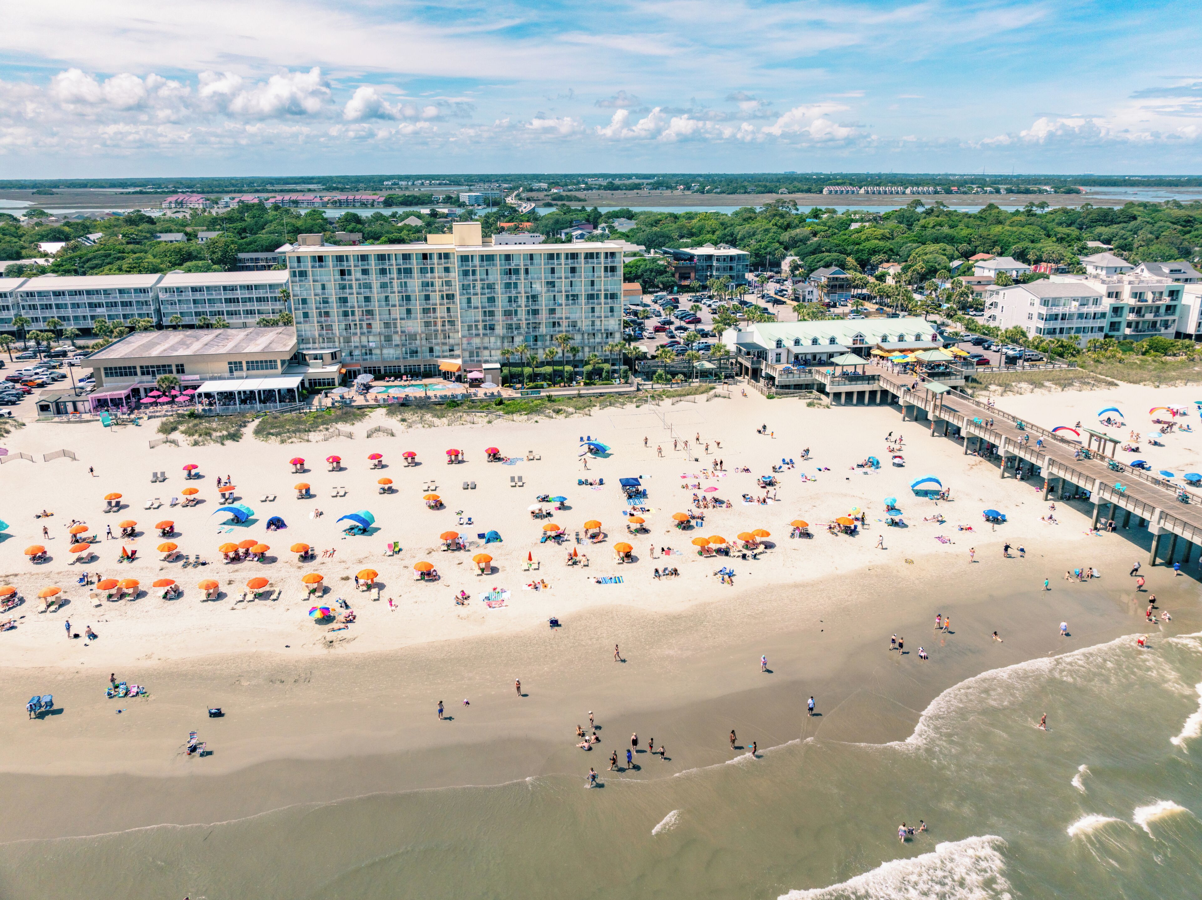 Une plage avec de nombreux parasols et beaucoup de monde, une jetée et des bâtiments en arrière-plan.