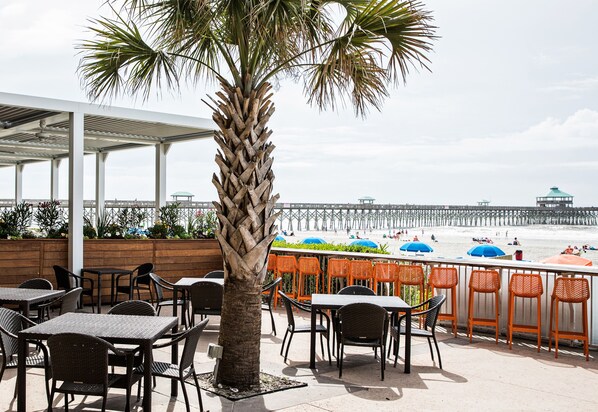 An outdoor seating area with tables and chairs, a palm tree, and a view of the beach and pier. - Tides Folly Beach, Charleston's Oceanfront Hotel (Folly Beach)