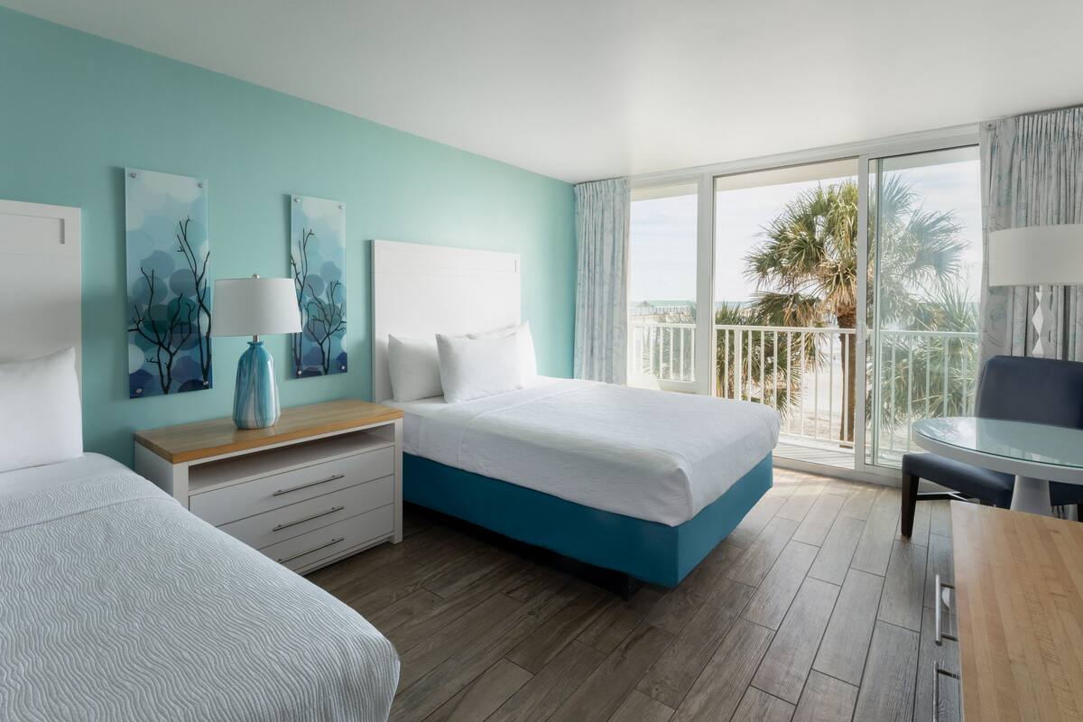 Une chambre d’hôtel avec un lit, un bureau, une chaise et un balcon donnant sur des palmiers.
