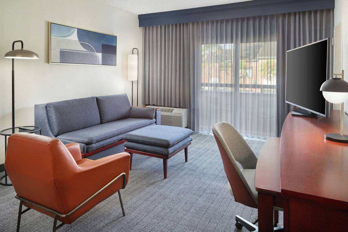 A hotel room with a grey sofa, a wooden desk with a TV, a red chair, and a lamp.