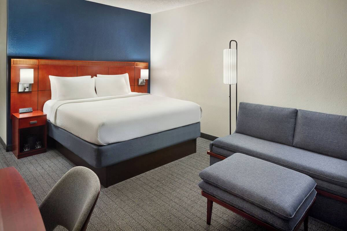 A hotel room with a bed, a sofa, a small table, and a nightstand with a lamp.