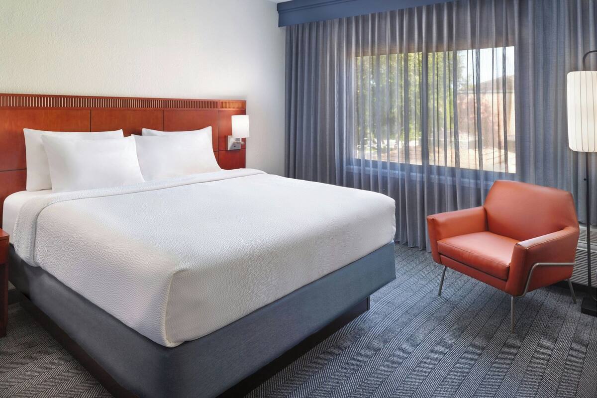 A hotel room with a large bed, a wooden headboard, a red armchair, and a window with curtains.