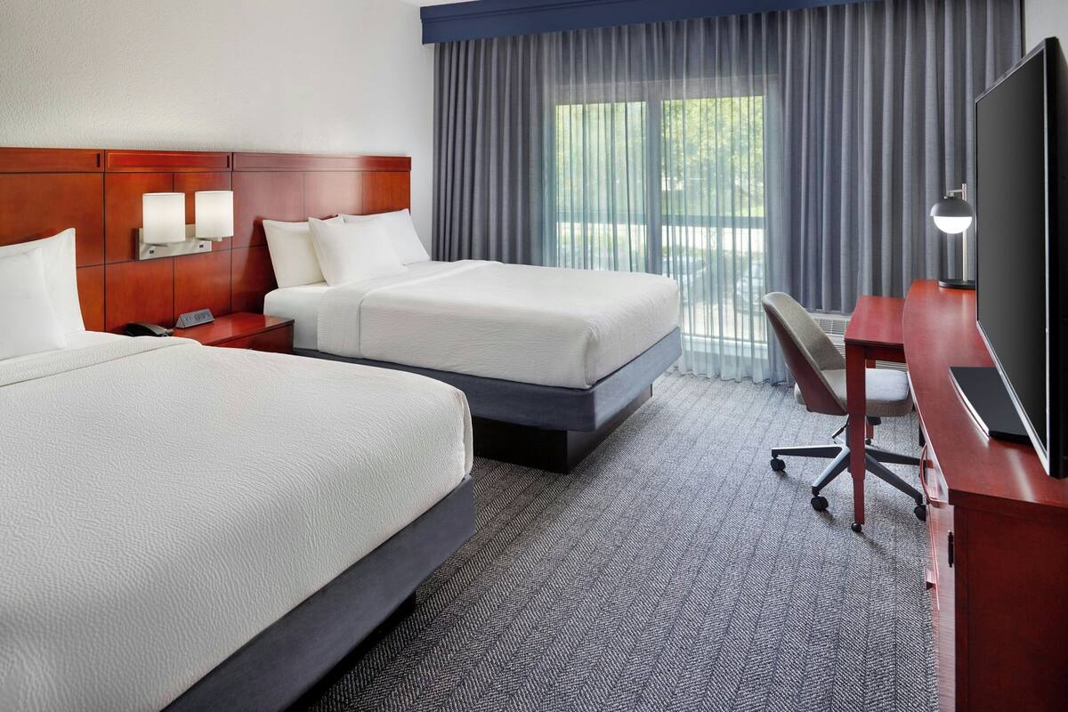 A hotel room with two beds, a desk, a TV, and a window with curtains.