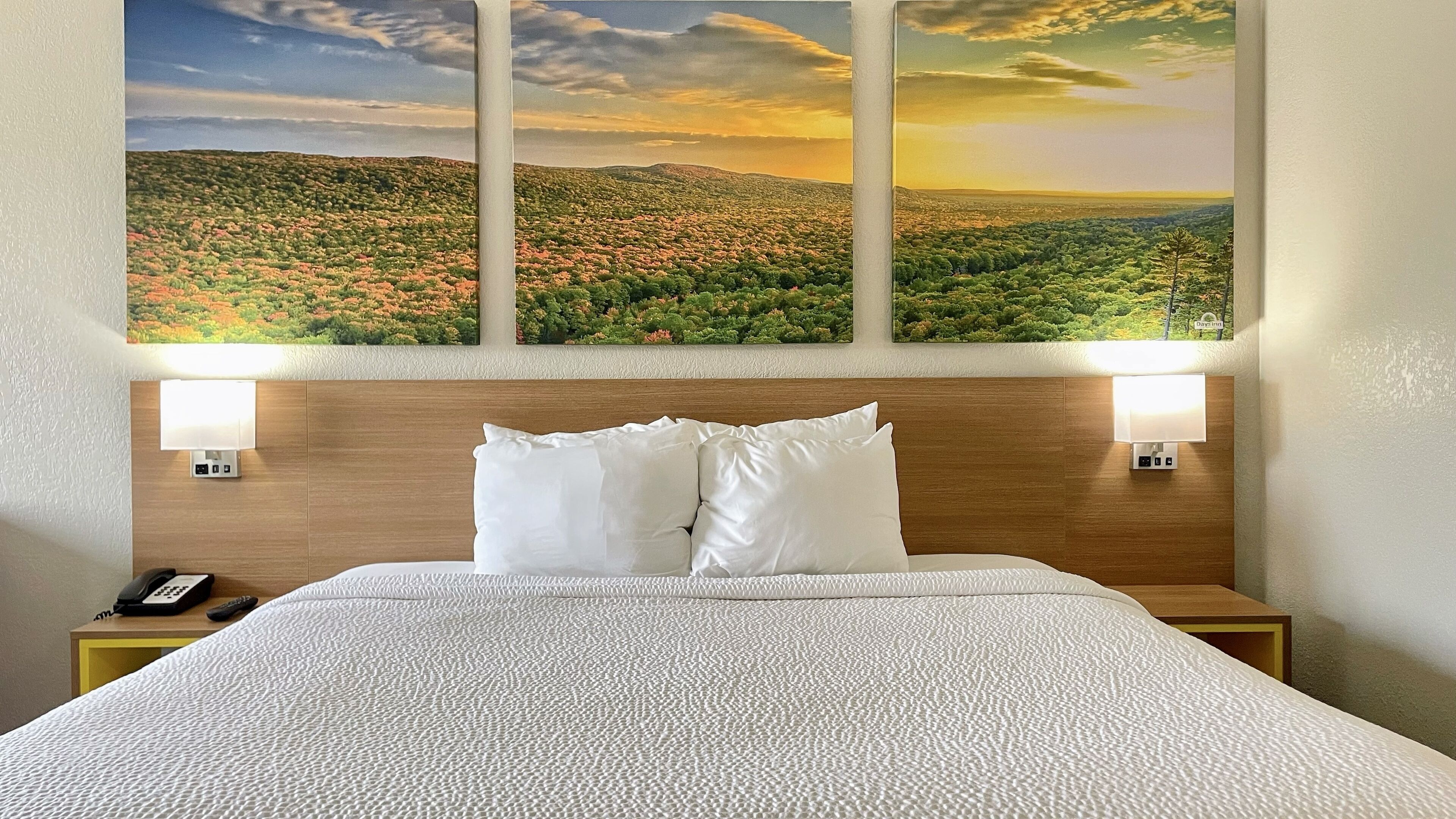 Ein Hotelzimmer mit einem großen Bett, zwei Nachttischlampen, einem hölzernen Kopfteil und einem an der Wand hängenden Landschaftsbild.