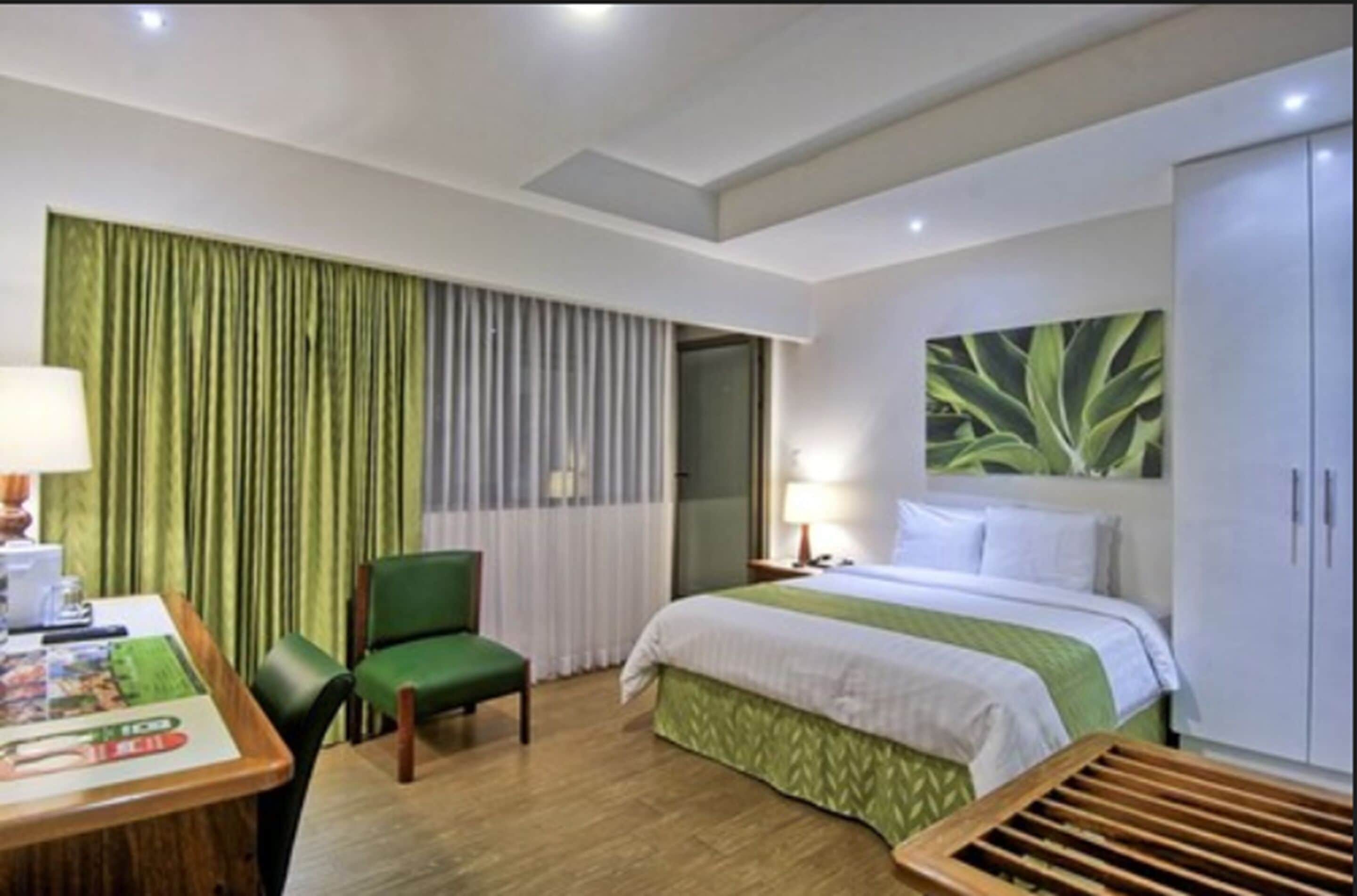 A hotel room with a bed, a desk with a chair, a window with curtains, and a wall-mounted artwork.