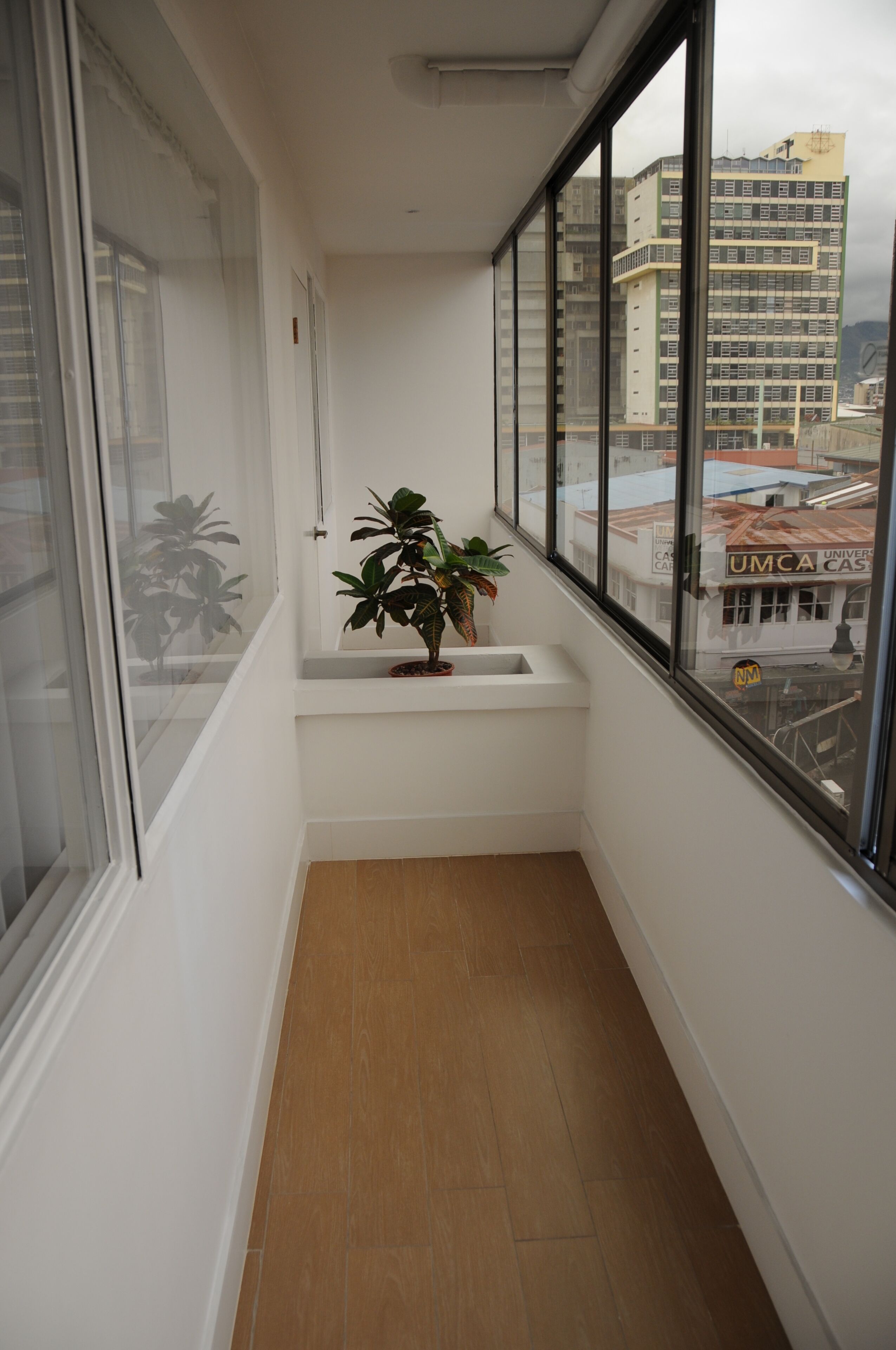 Un balcón estrecho con una maceta, un banco blanco y vistas a edificios a través de amplias ventanas.