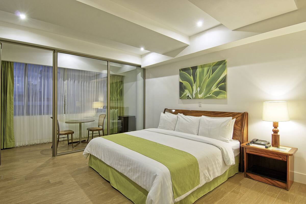 A hotel room with a bed, a bedside table with a lamp, a chair, a small table, and a sliding glass door leading to a balcony.