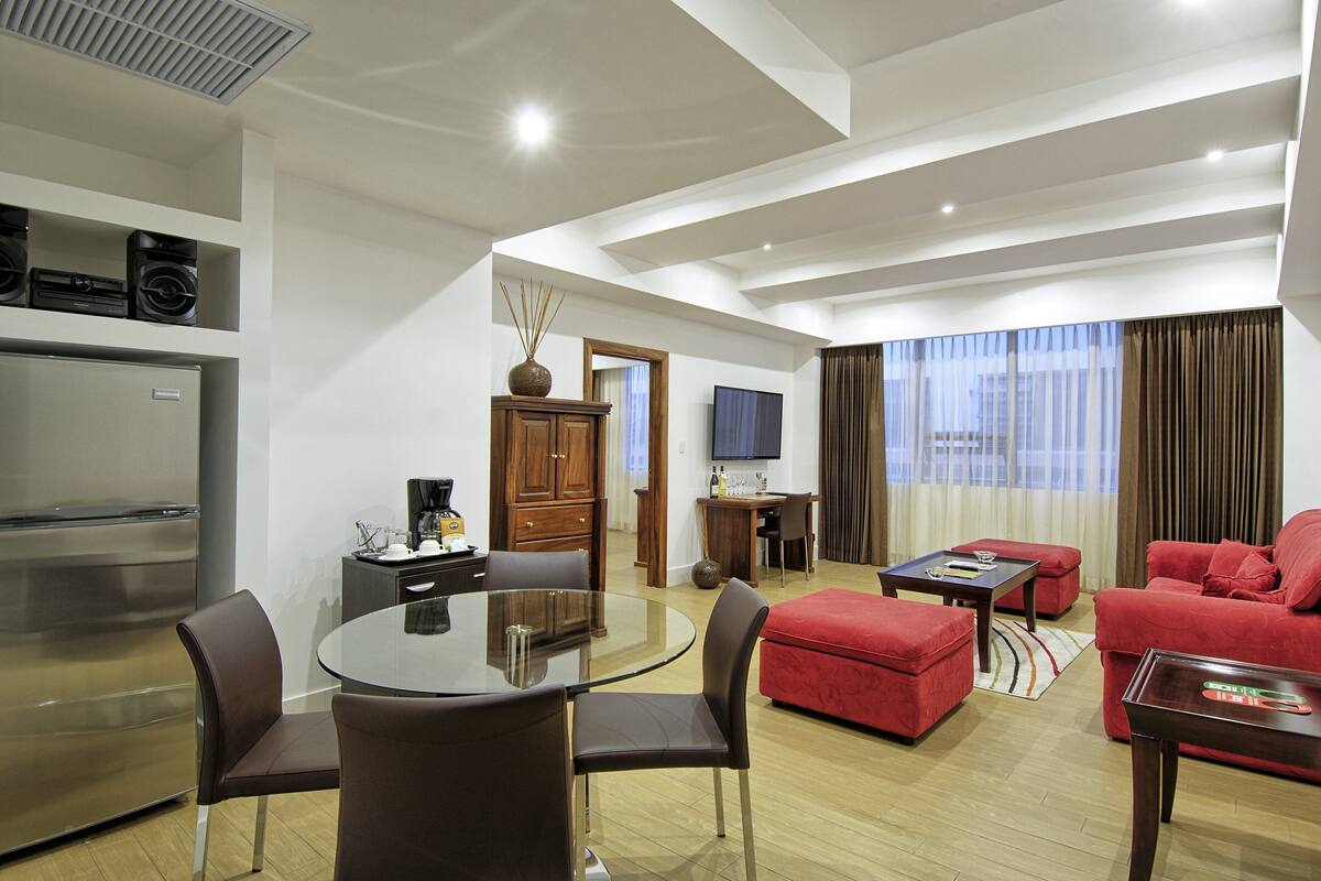 A modern living room with a glass dining table, red sofas, a flat-screen TV, and a kitchen area with a refrigerator and microwave.