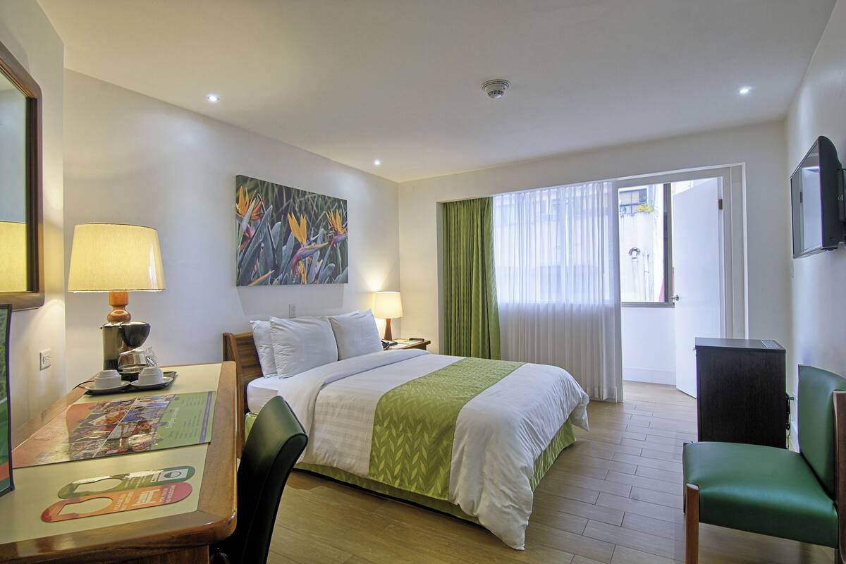 A hotel room with a bed, a desk with a lamp, a chair, a television, and a painting on the wall.
