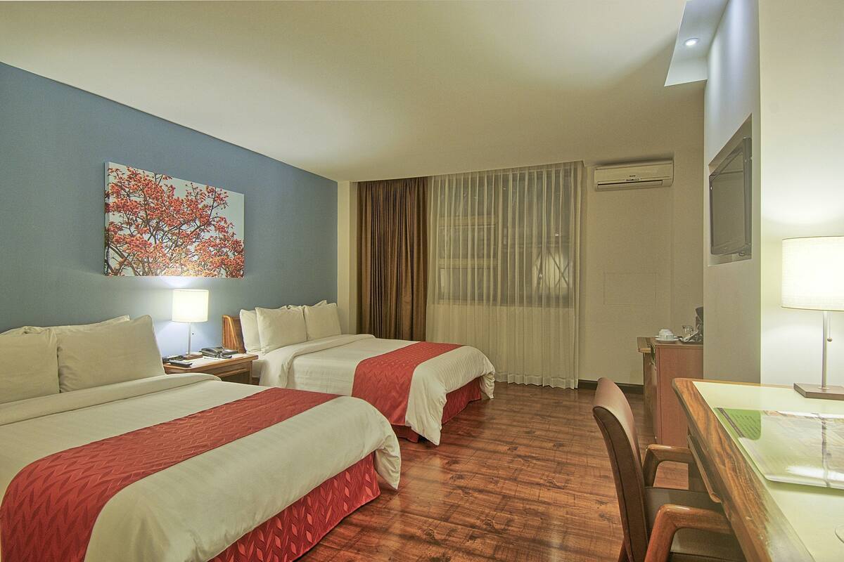 A hotel room with two beds, a desk, a chair, a lamp, and a painting on the wall.