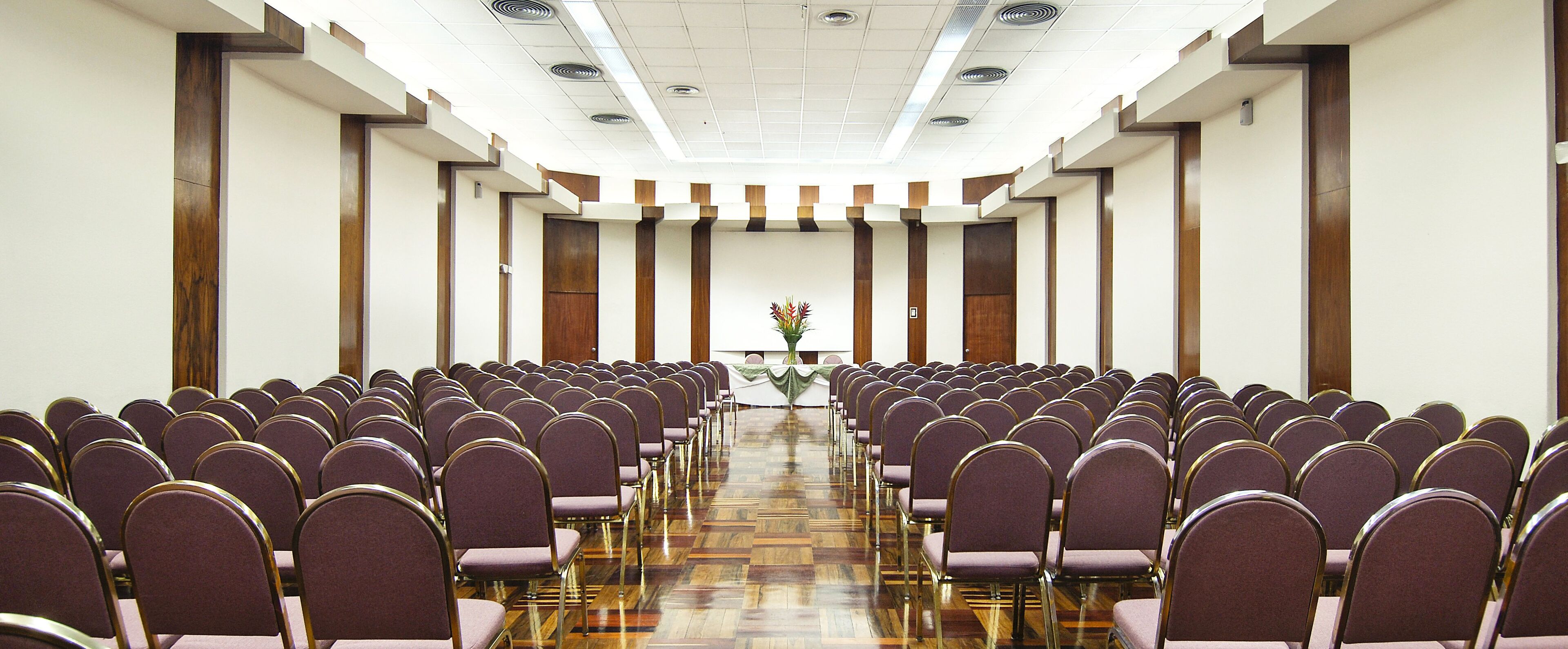 A conference room with rows of chairs facing a podium, wooden paneling on the walls, and a polished wooden floor.