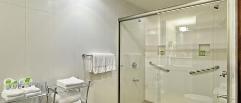 A modern bathroom with a glass-enclosed shower, white tiled walls, and a glass shelf holding folded towels.