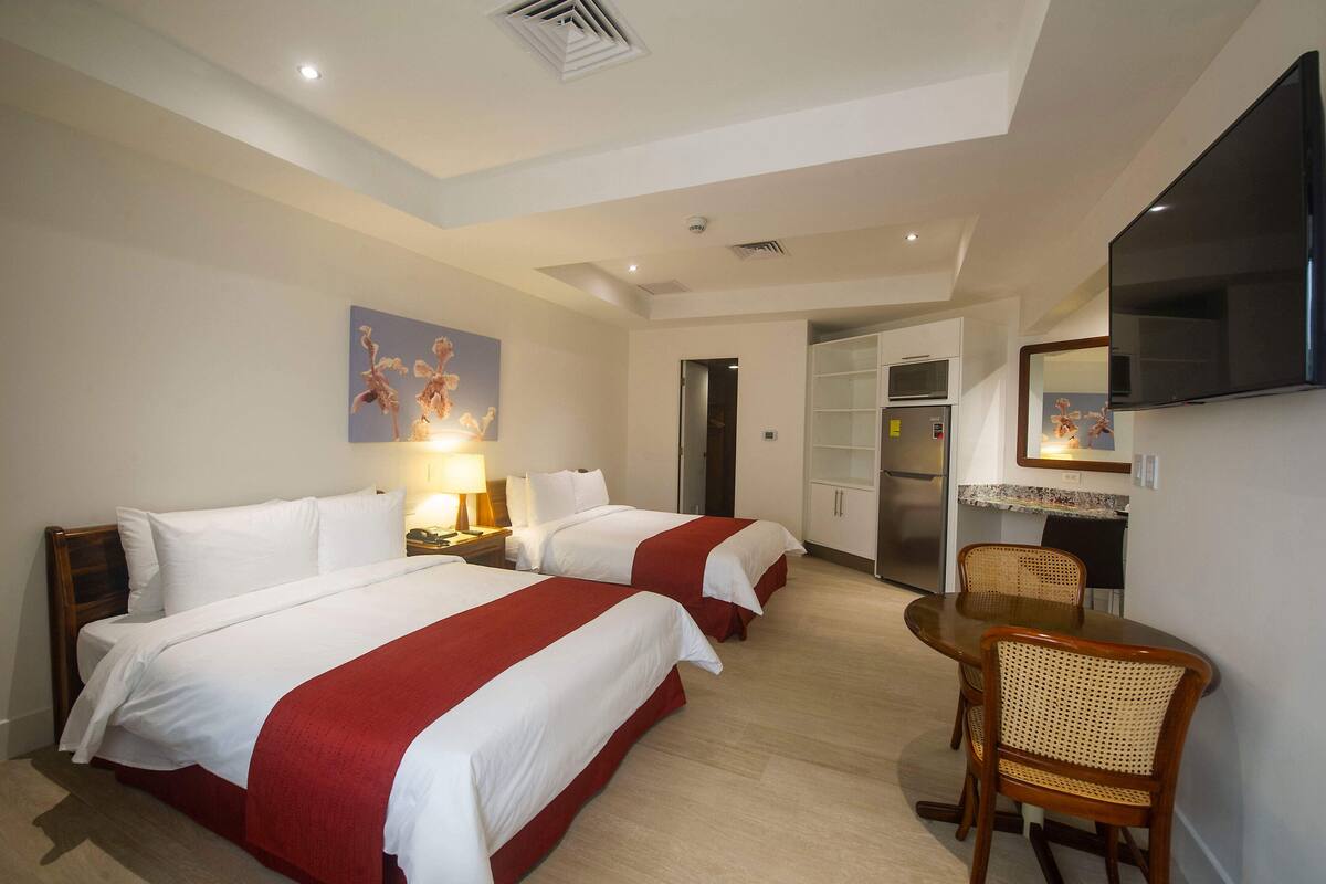 A hotel room with two beds, a dining table with chairs, a TV mounted on the wall, and a kitchen area visible through an open door.