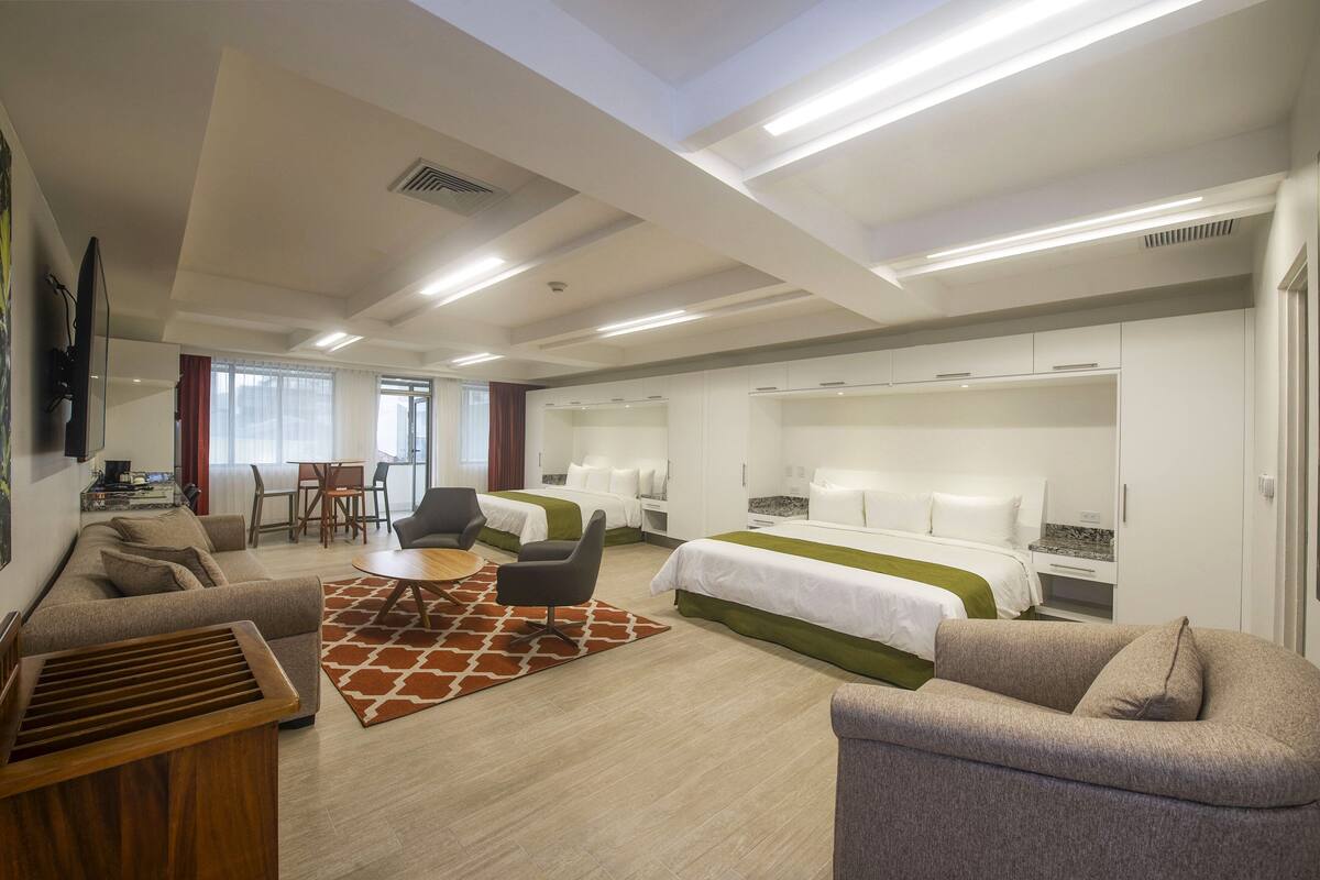 A modern hotel room with a sofa, two beds, a dining table, and a TV.