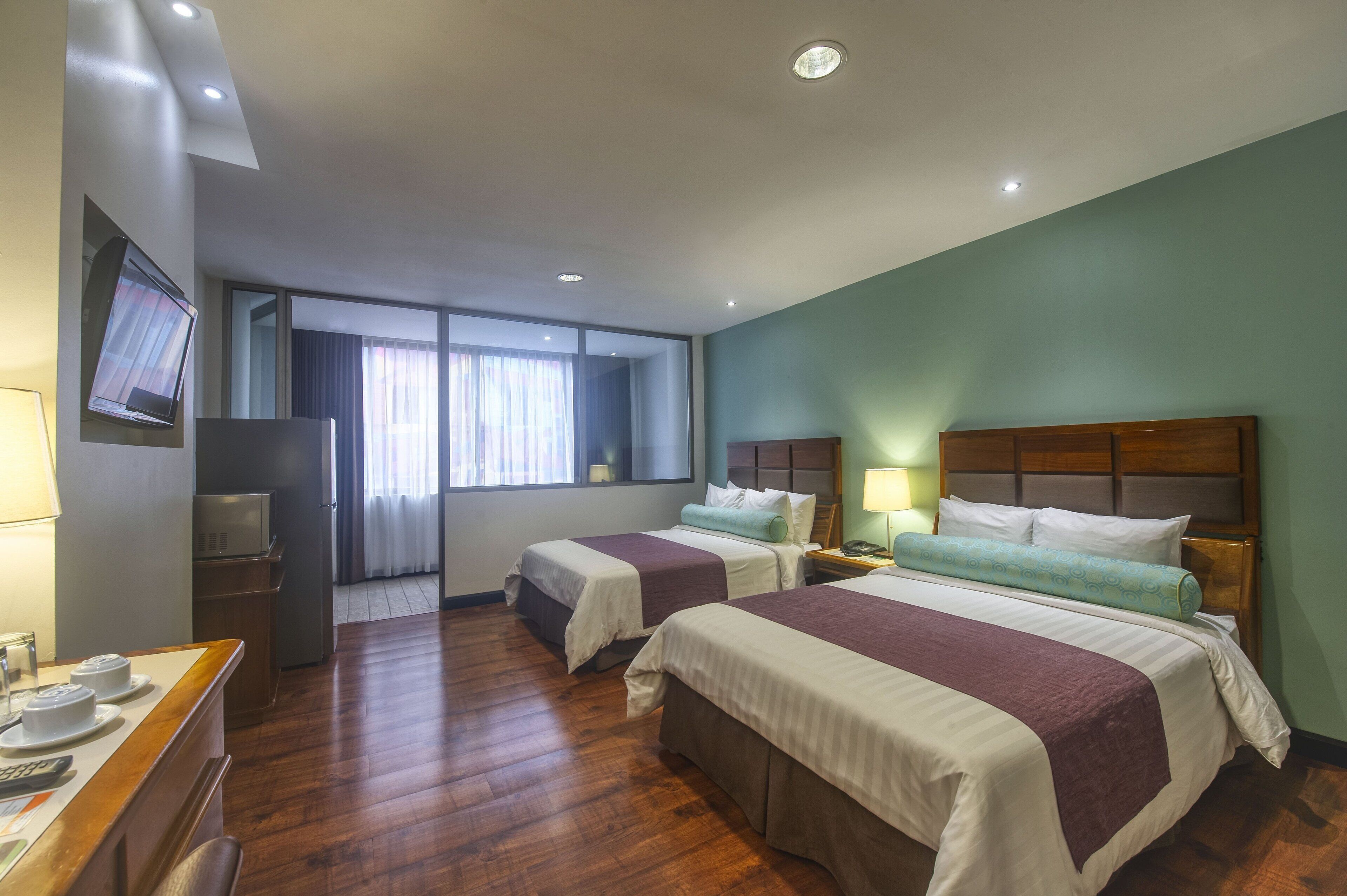 Habitación de hotel con dos camas, televisión, escritorio con lámpara y ventana con cortinas.