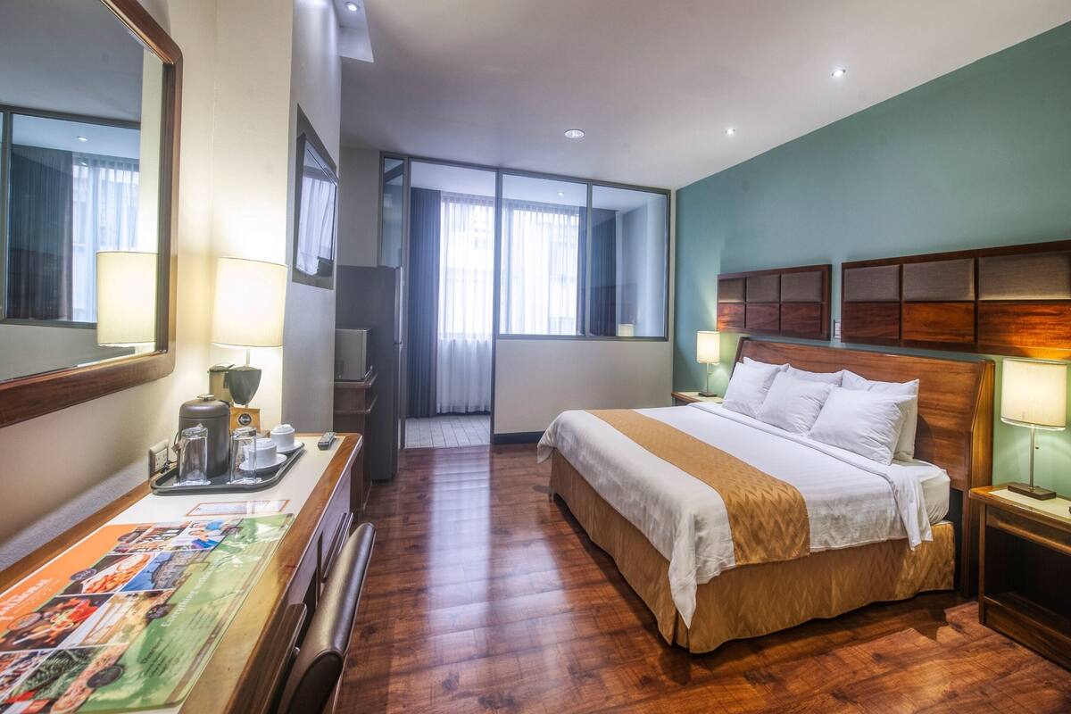 A hotel room with a wooden bed, a desk with a coffee maker, a mirror, and a window with curtains.
