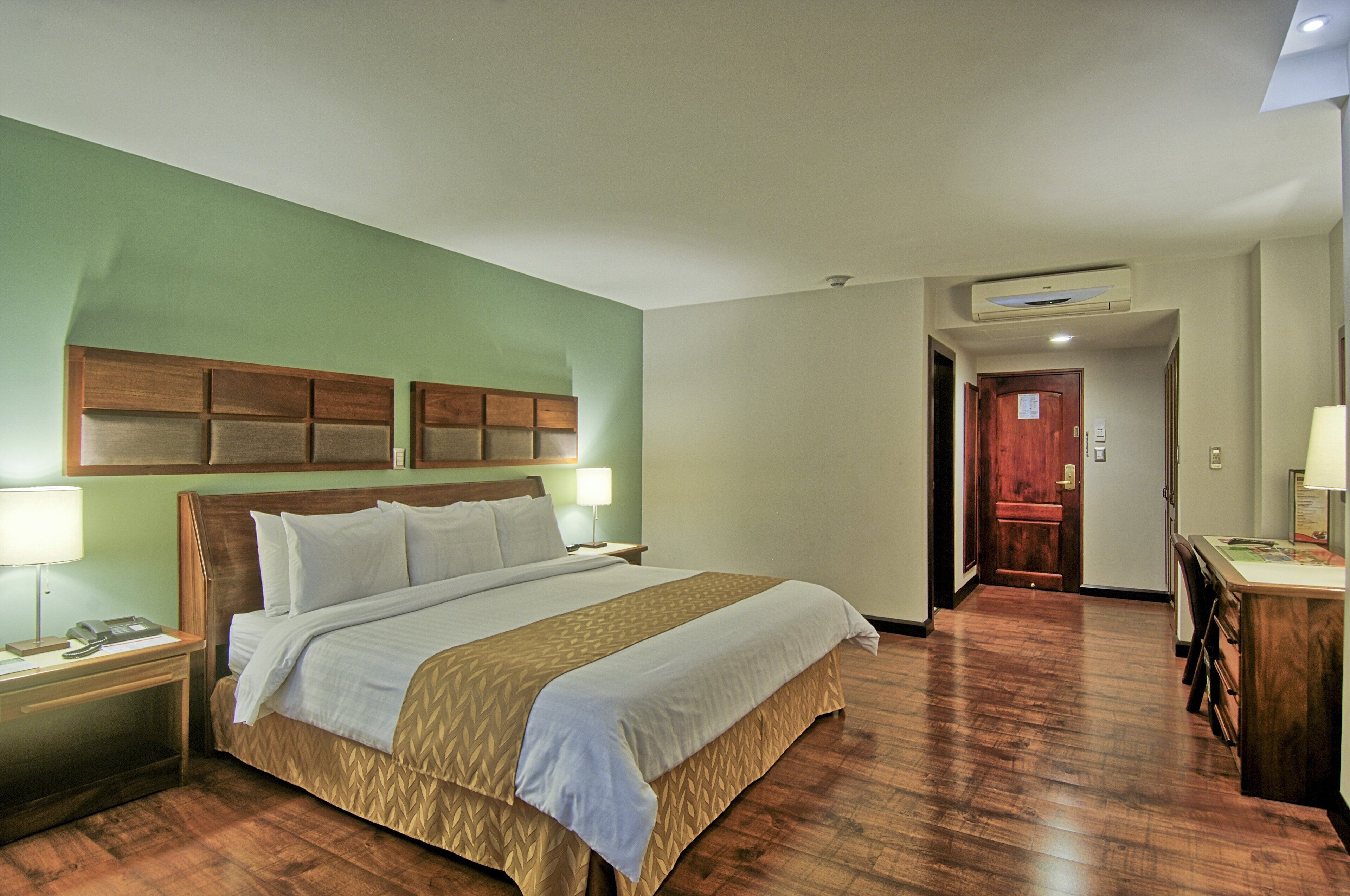 A hotel room with a large bed, wooden headboard, bedside tables with lamps, a desk with a chair, and a door leading to another room.