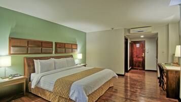Una habitación de hotel con una cama grande, cabecero de madera, mesitas de noche con lámparas, un escritorio con una silla y una puerta que da a otra habitación.