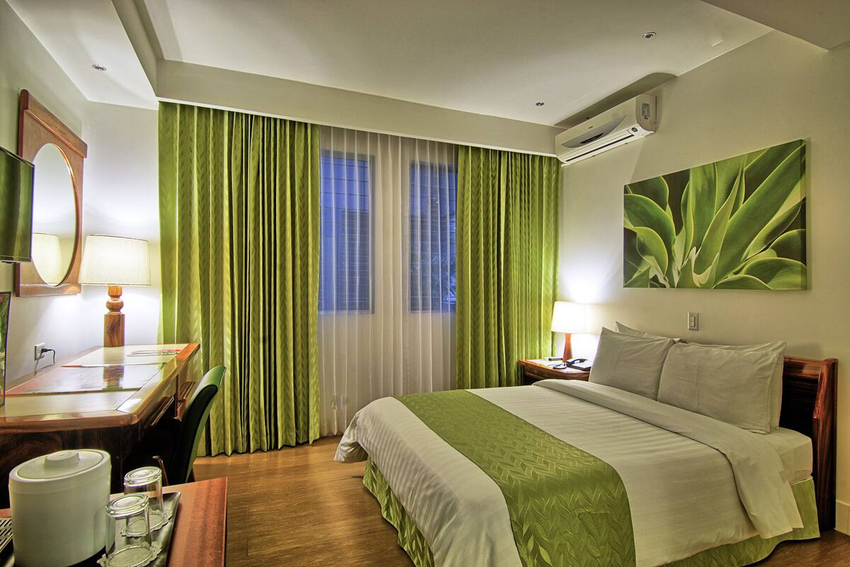 A hotel room with a bed, a desk, a chair, a lamp, a mirror, and a painting.