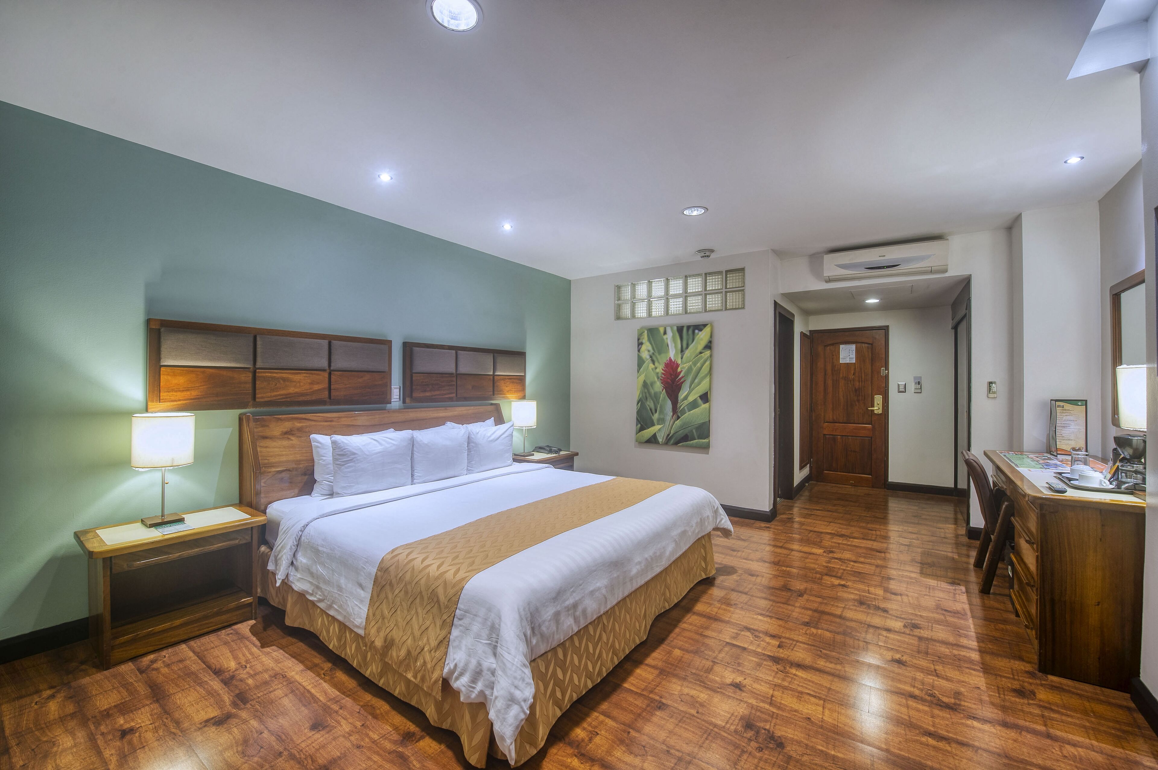 A hotel room with a wooden bed, a desk, and a chair. There is a door leading to another room and an air conditioning unit on the wall.