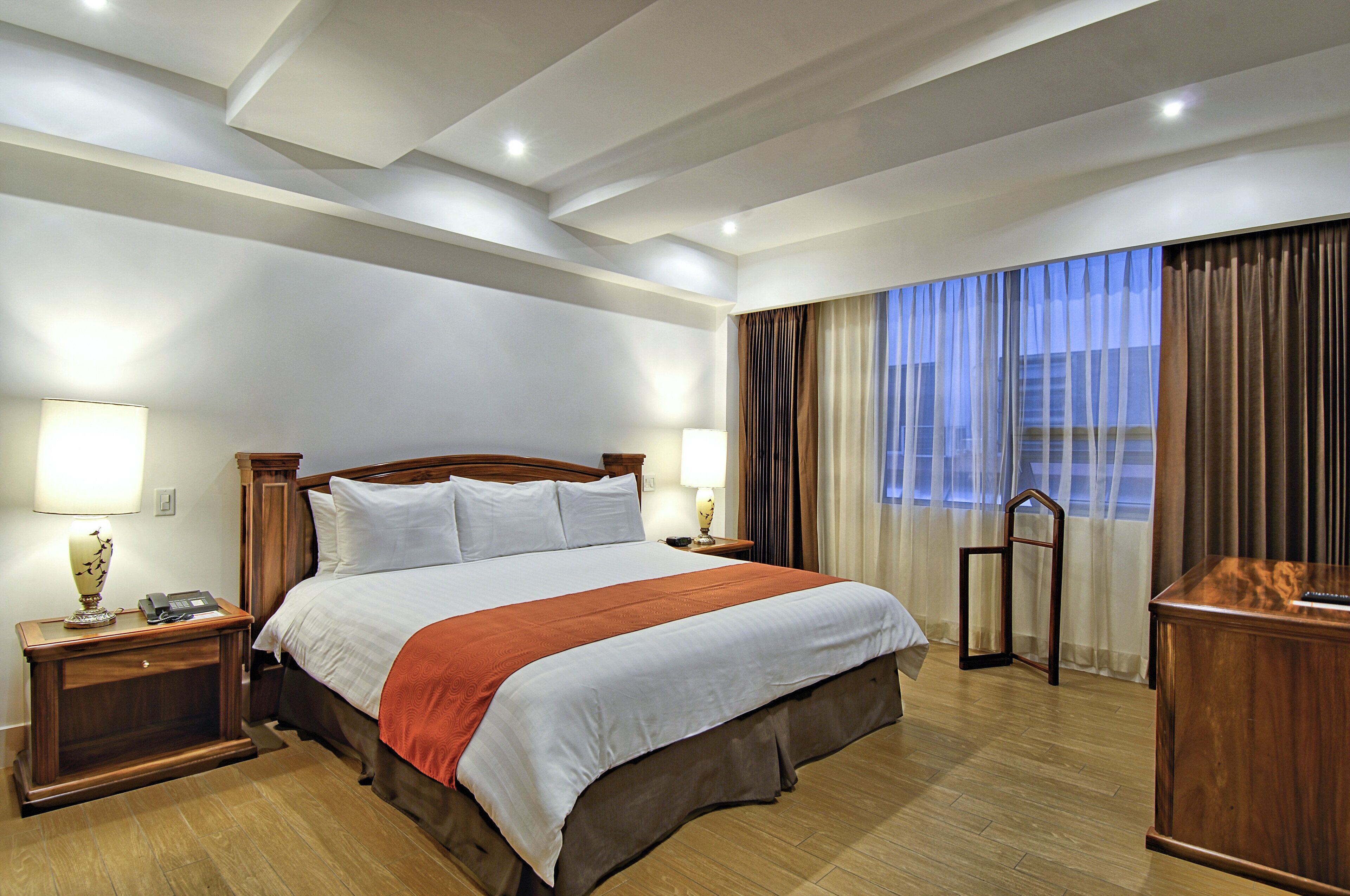 A hotel room with a large bed, wooden headboard, two bedside tables with lamps, a desk with a chair, and a view of the city through the window.