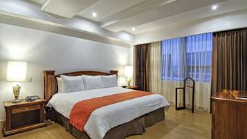 A hotel room with a large bed, wooden headboard, two bedside tables with lamps, a desk with a chair, and a view of the city through the window.