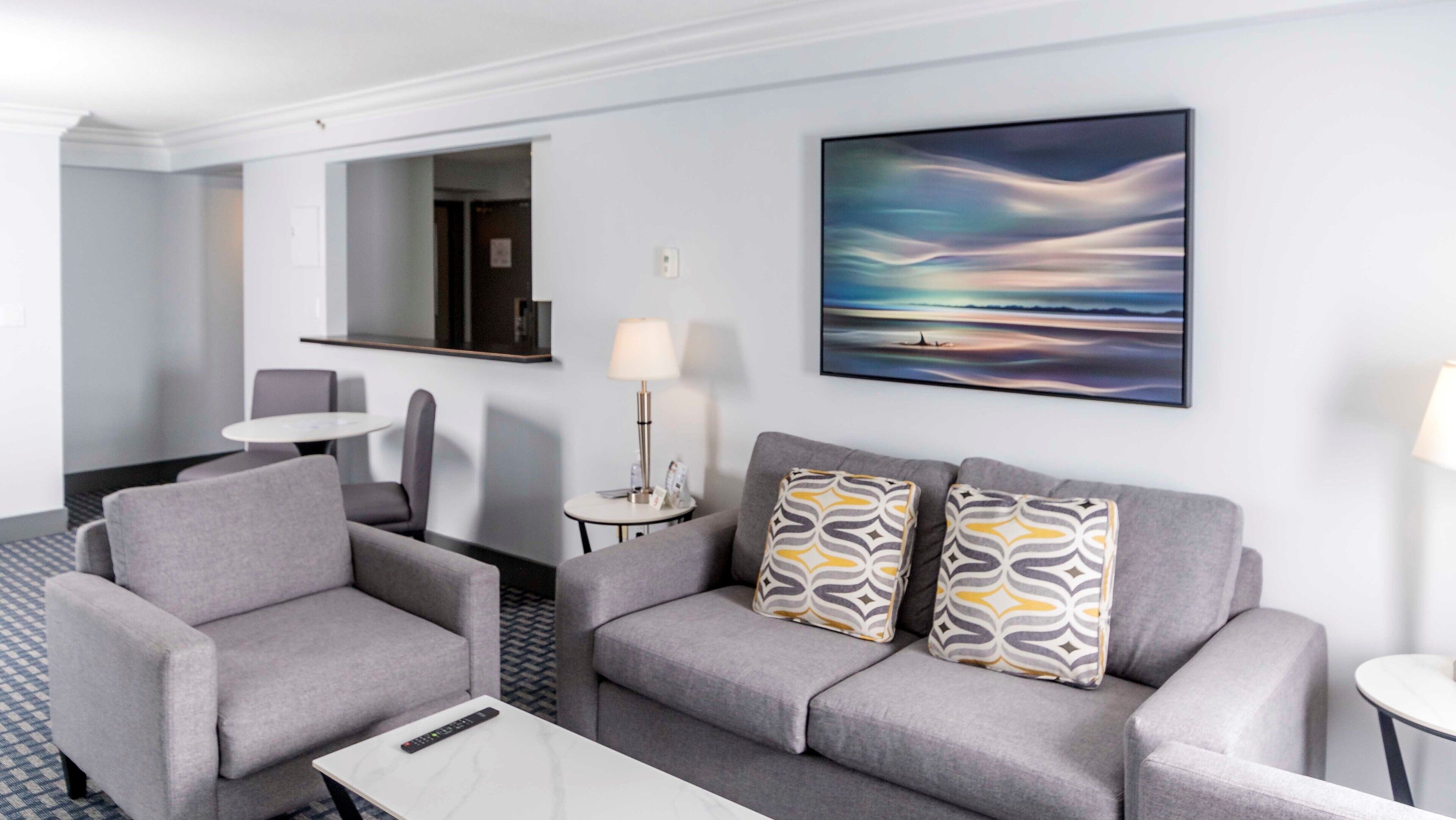A modern hotel room with a grey sofa set, a small round table, a coffee table, a lamp, and a painting of a landscape.