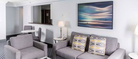 A modern hotel room with a grey sofa set, a small round table, a coffee table, a lamp, and a painting of a landscape.