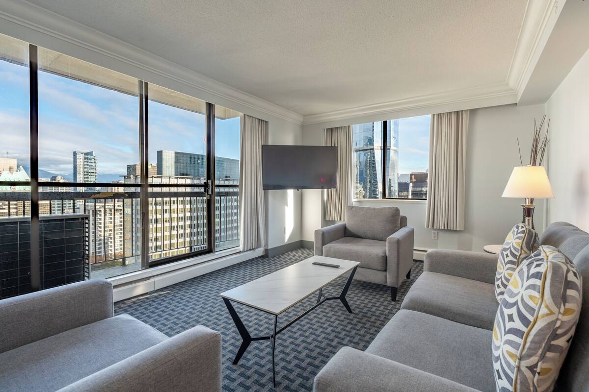 A modern hotel room with a large window, a flat-screen TV, a sofa, a coffee table, and a lamp.