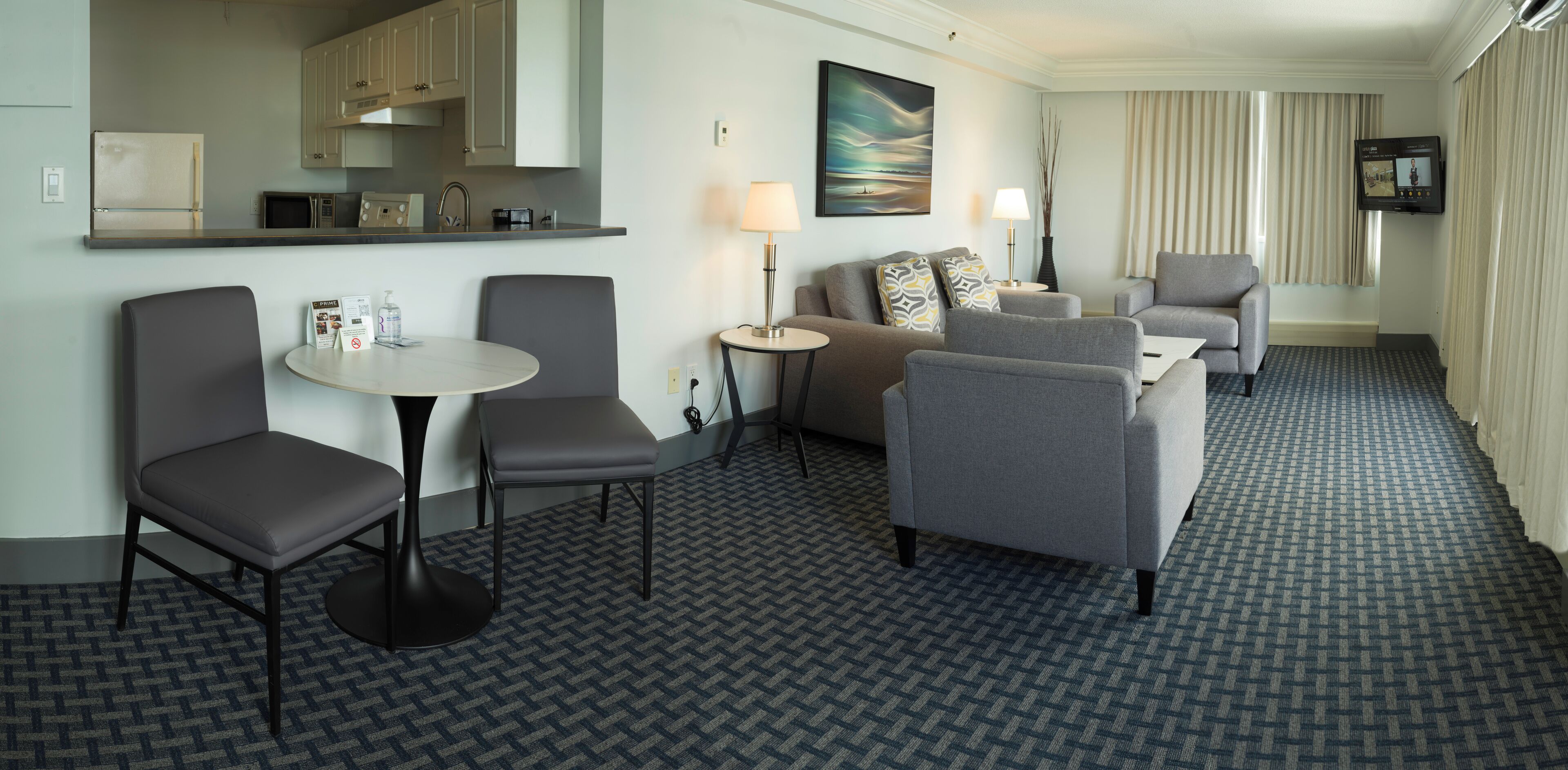 Une chambre d’hôtel avec une kitchenette, un coin repas comprenant une table ronde et deux chaises, un canapé, une télévision et une moquette à motifs.