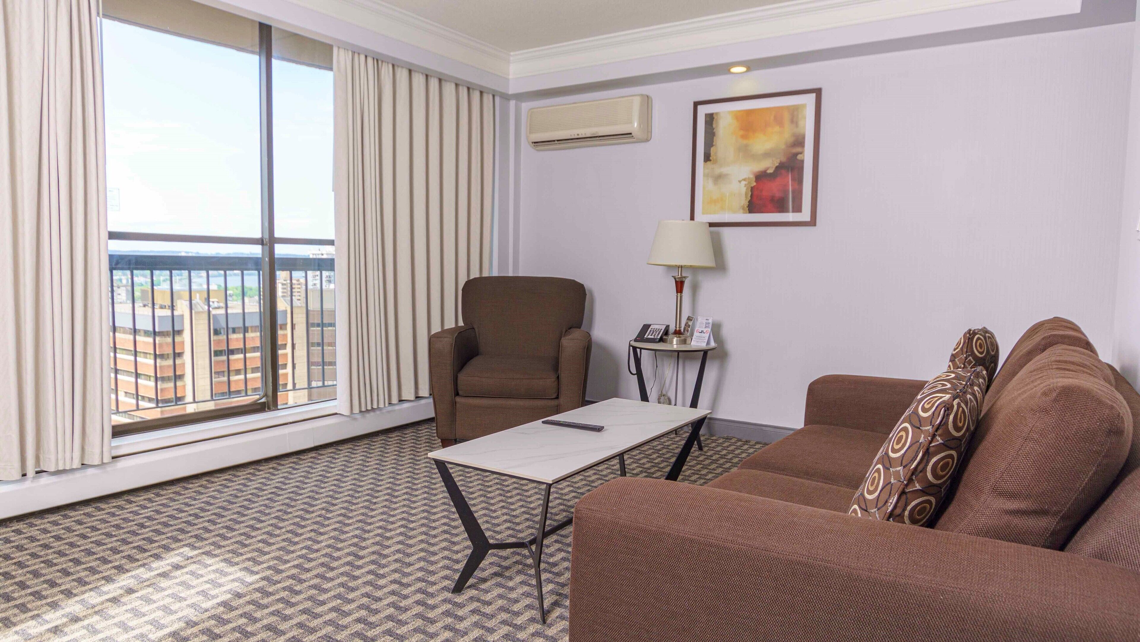 Une chambre d’hôtel avec une grande fenêtre, un fauteuil marron, un canapé, une petite table et une lampe.