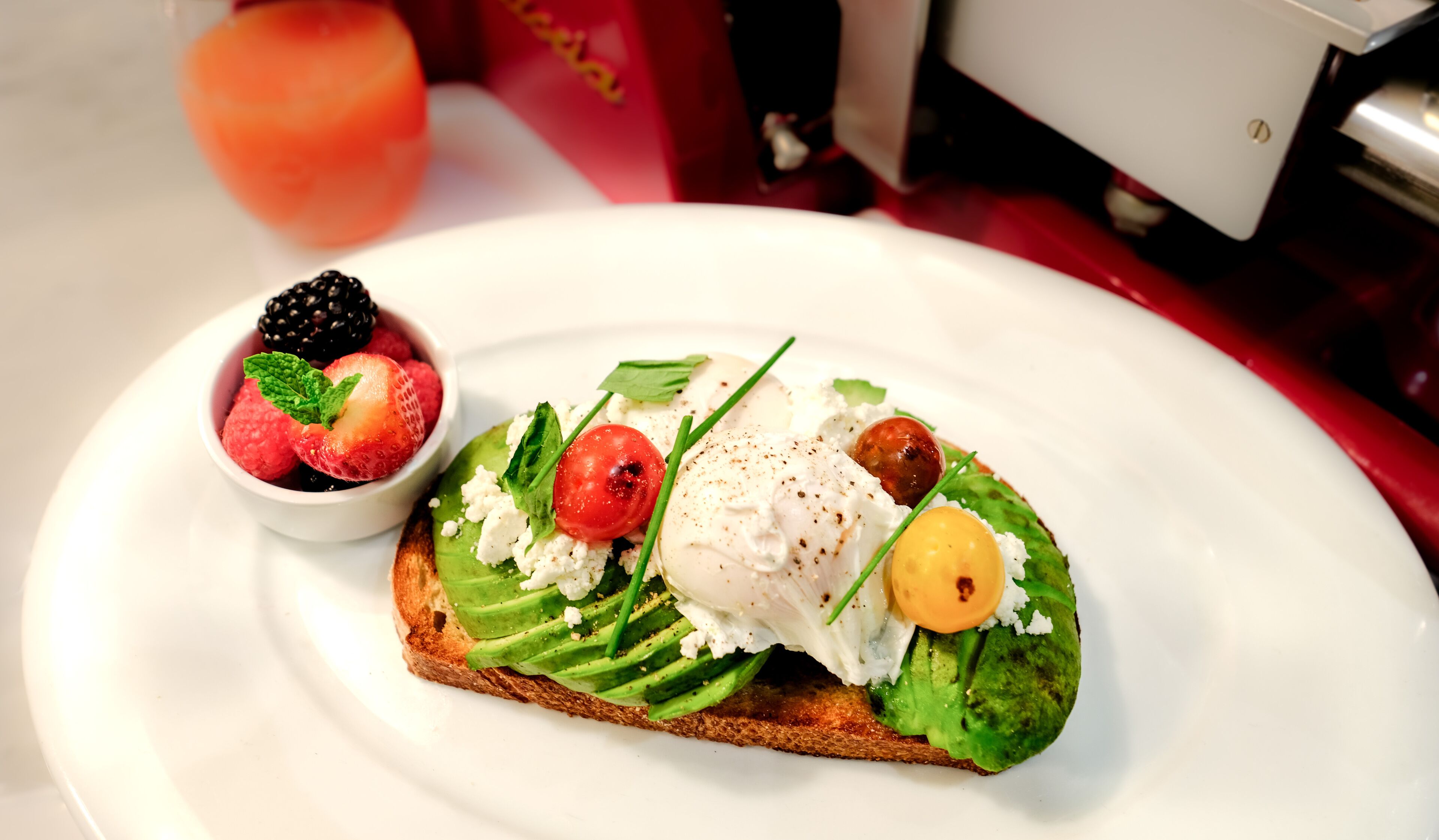 Une assiette avec un sandwich garni d’avocat, de tomates et d’un œuf poché, accompagnée d’un bol de fruits rouges variés.