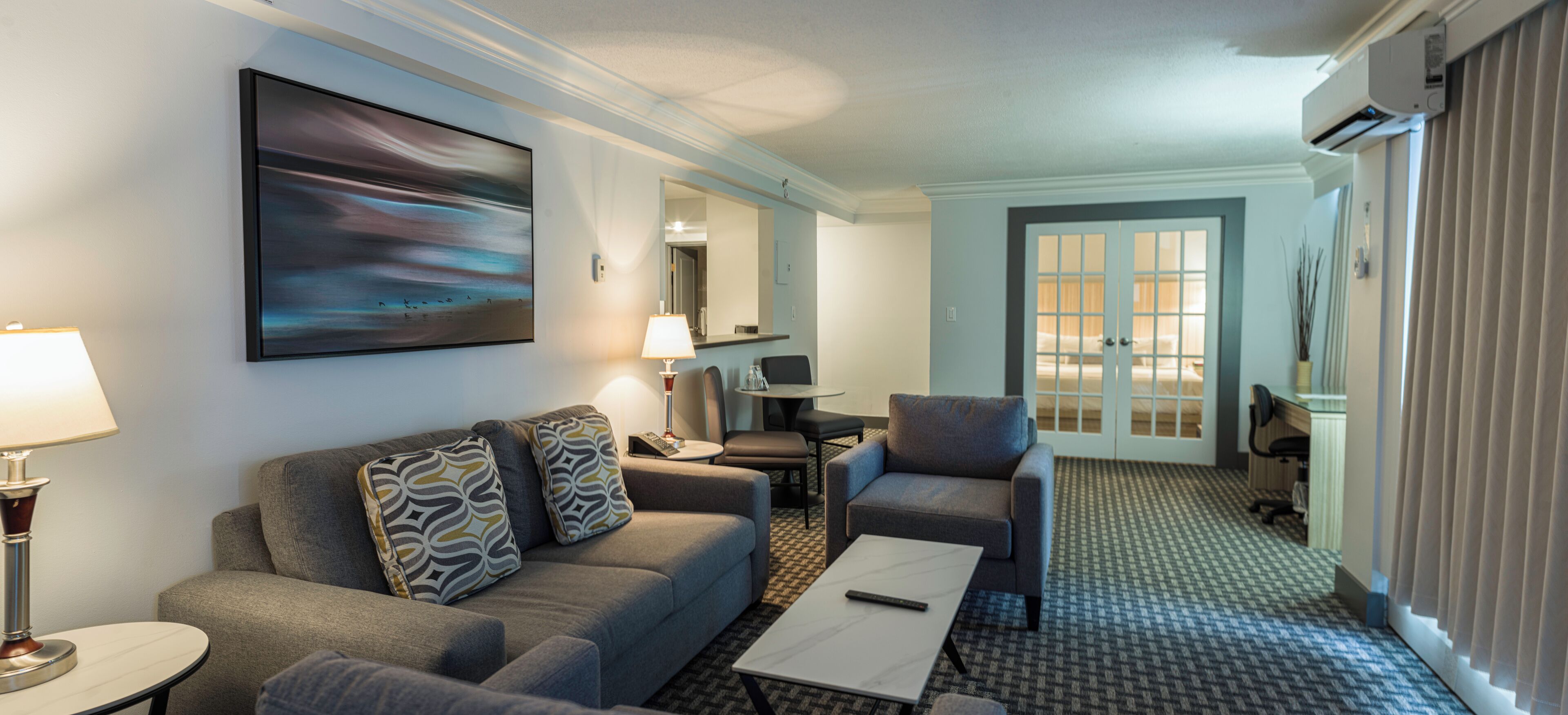 A modern hotel room with a grey sofa, armchairs, a coffee table, and a large painting on the wall.