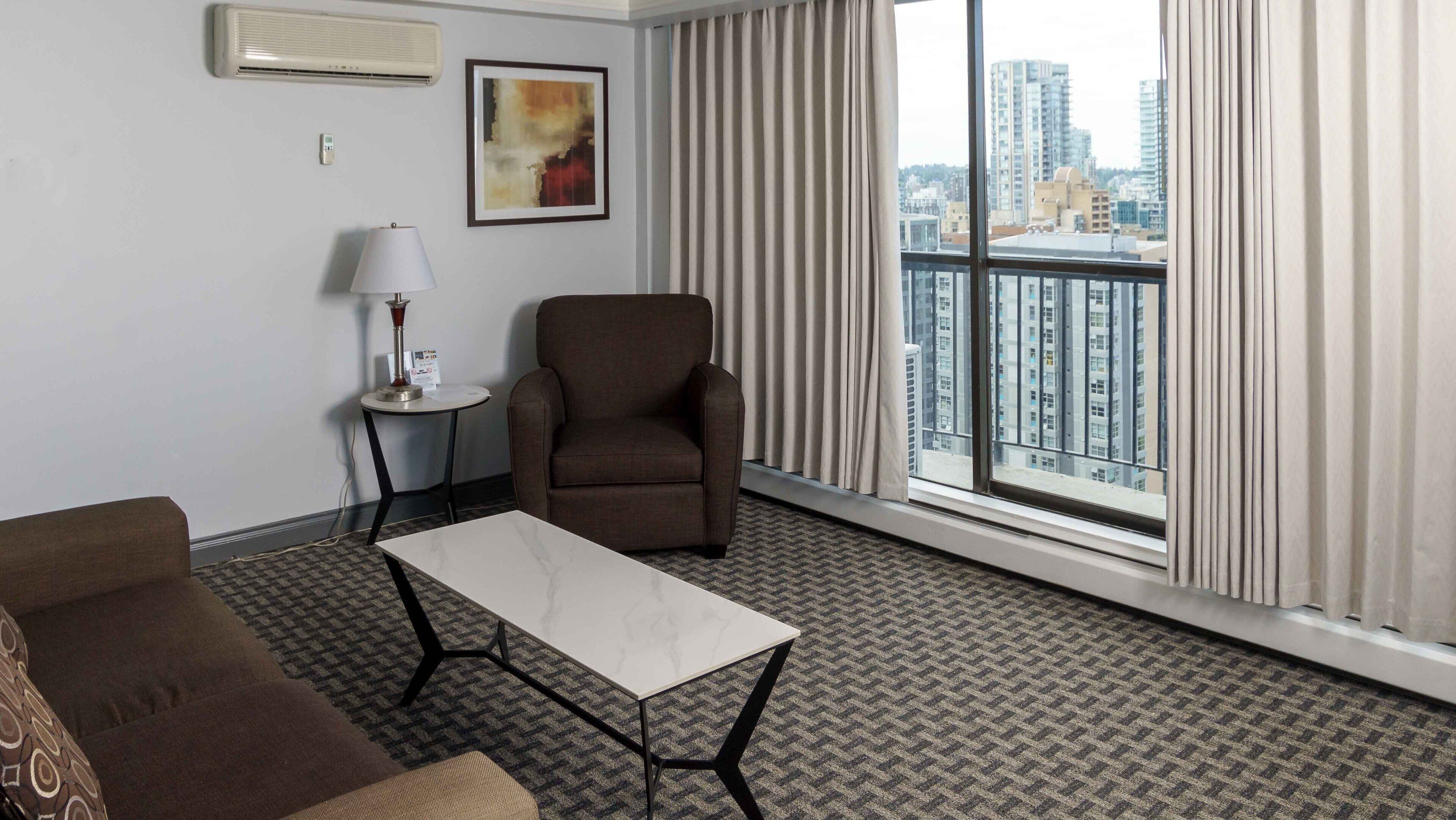 Habitación de hotel con sofá, una mesita, una silla, lámpara, un cuadro y un ventanal con vistas a la ciudad.