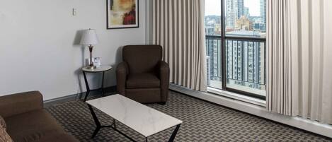 Habitación de hotel con sofá, una mesita, una silla, lámpara, un cuadro y un ventanal con vistas a la ciudad.