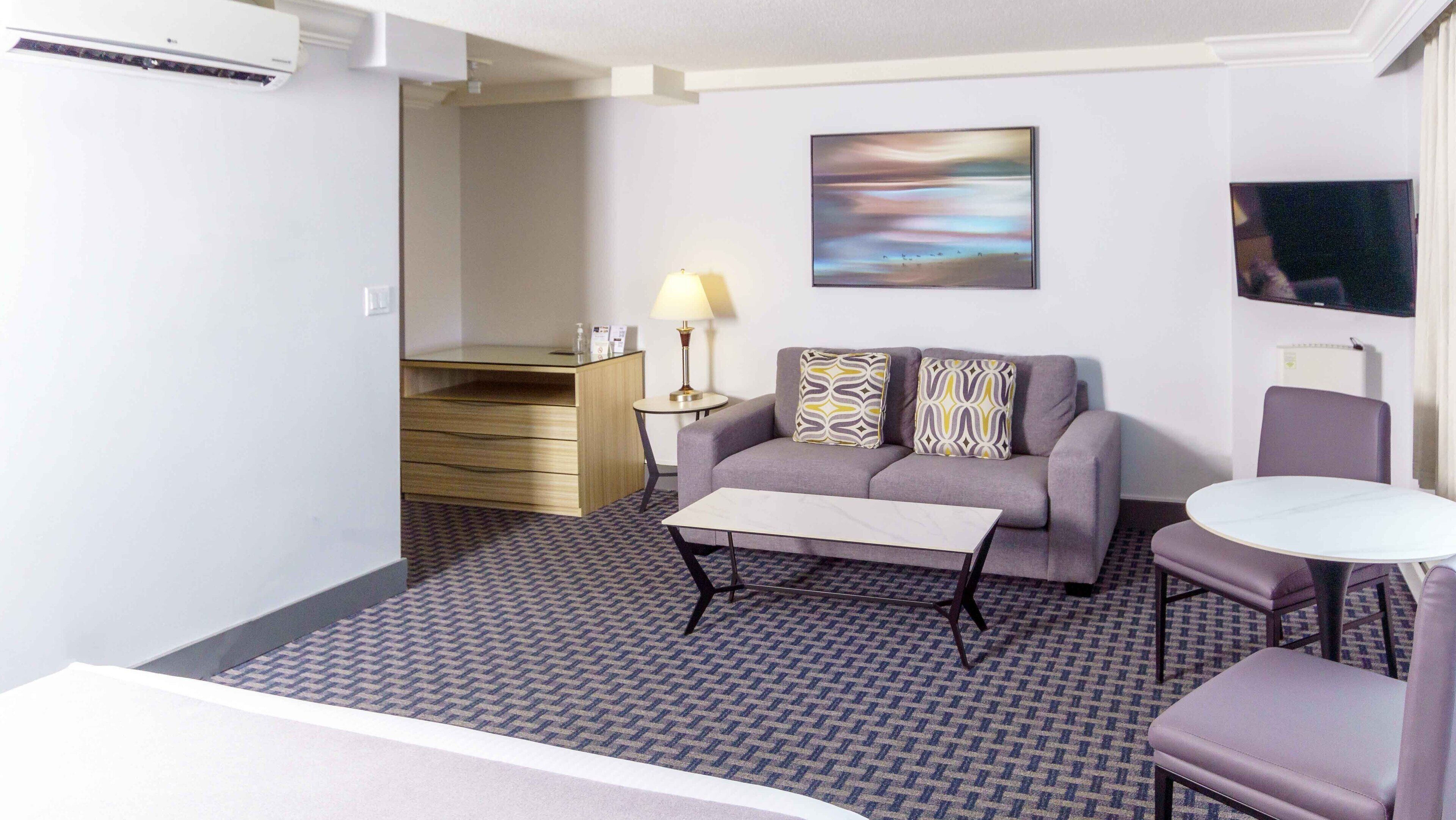 Habitación de hotel con sofá, una mesita, lámpara, un cuadro en la pared y televisión.