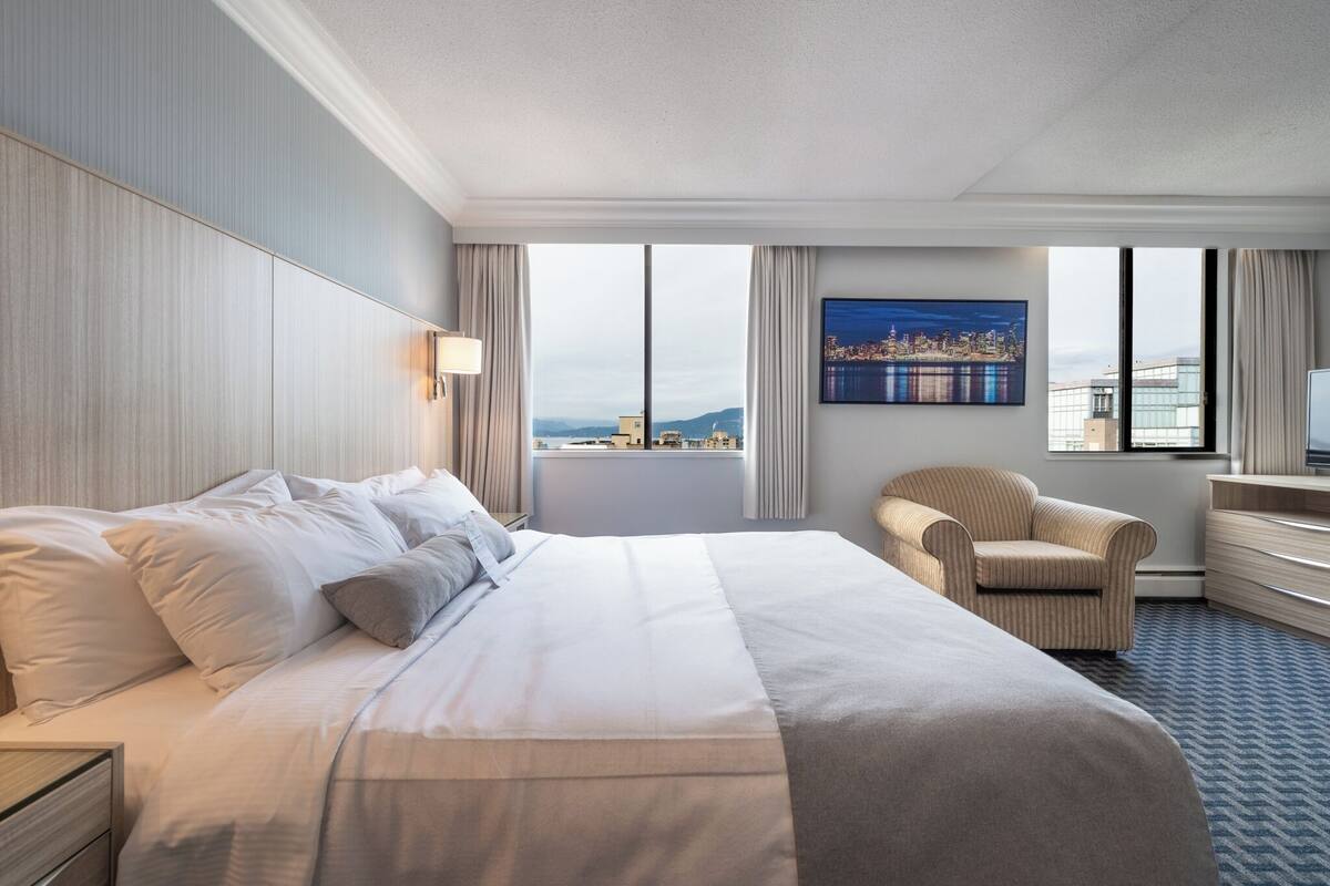A hotel room with a large bed, a chair, a desk, and a cityscape view through the windows.