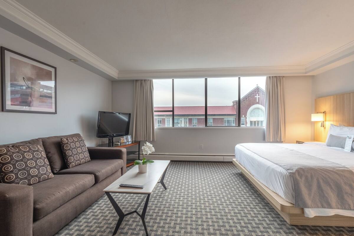 A hotel room with a bed, a sofa, a TV, and a window with a view of a building.