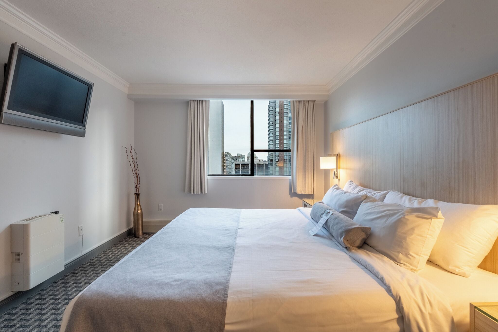A hotel room with a large bed, a flat-screen TV mounted on the wall, a window with curtains, and a view of the cityscape.
