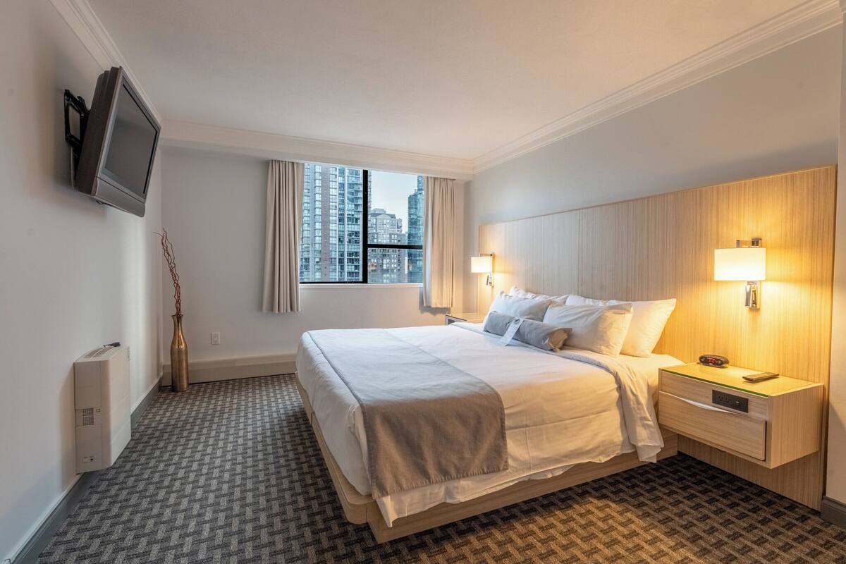 A hotel room with a large bed, a flat-screen TV mounted on the wall, a bedside table with a lamp, and a view of the city through the window.