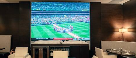 Habitación de hotel con una amplia pantalla de televisión mostrando un evento deportivo, una mesa de comedor y disposición de asientos.